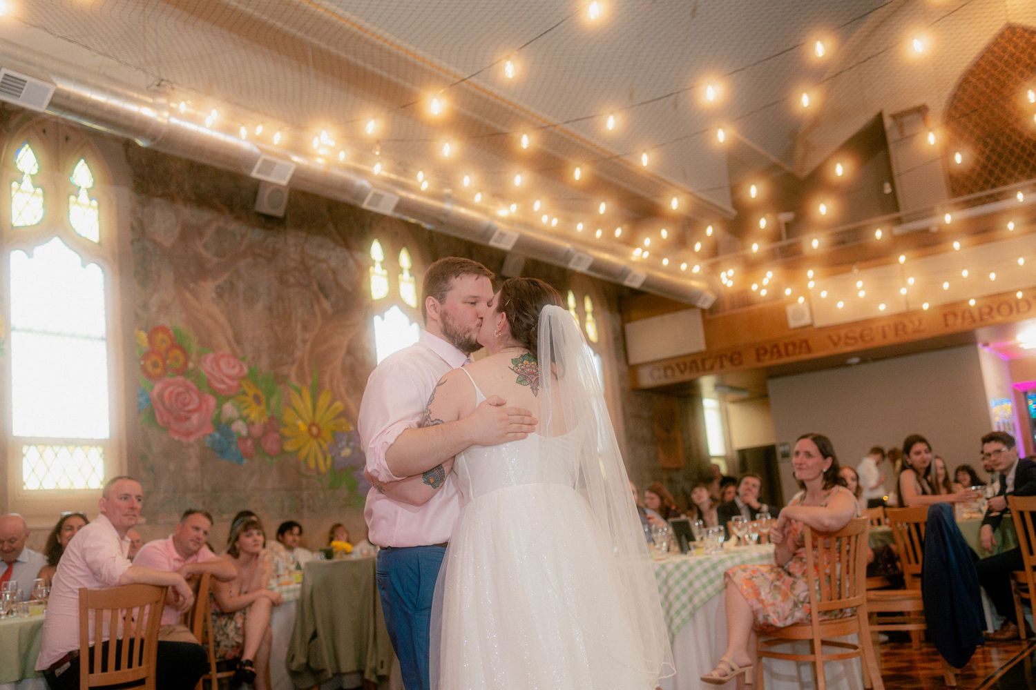 Bride and groom share first dance under string lights in elegant wedding reception hall with floral mural backdrop.