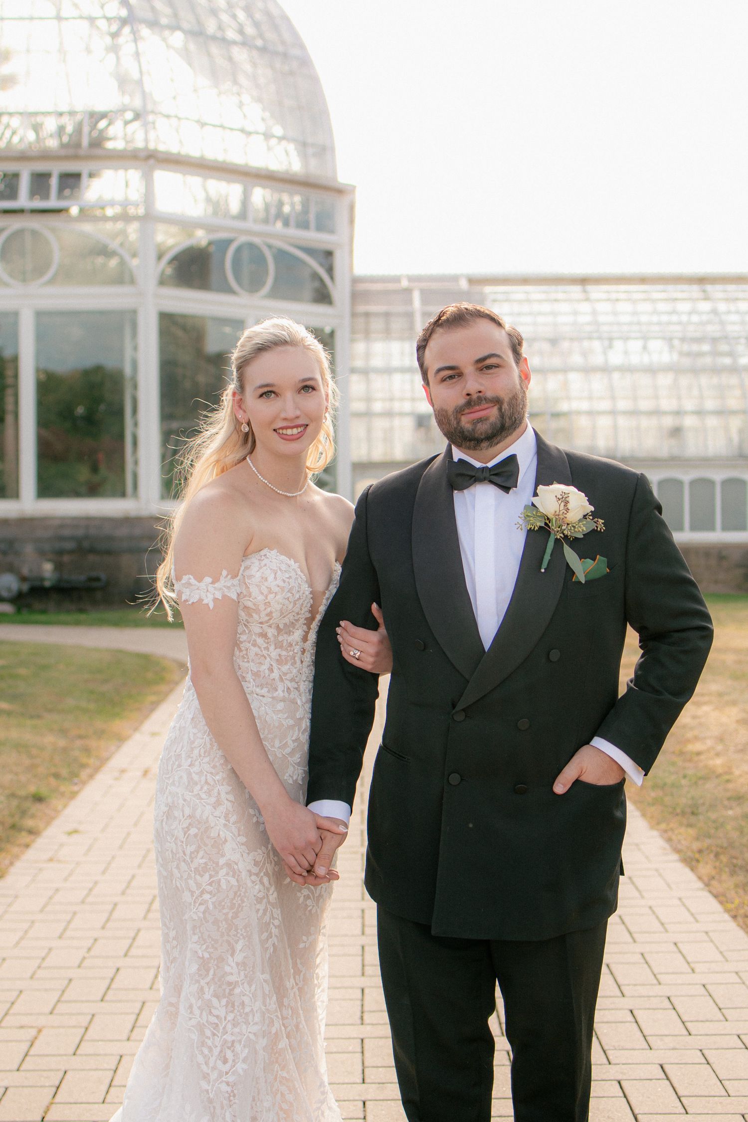 Elegant bride in lace gown and groom in black tuxedo pose together outside a stunning glass greenhouse.