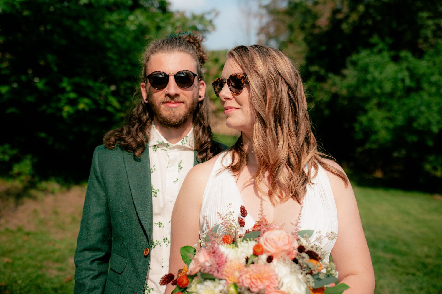 Bride and groom wearing sunglasses pose romantically outdoors with lush green trees in background.
