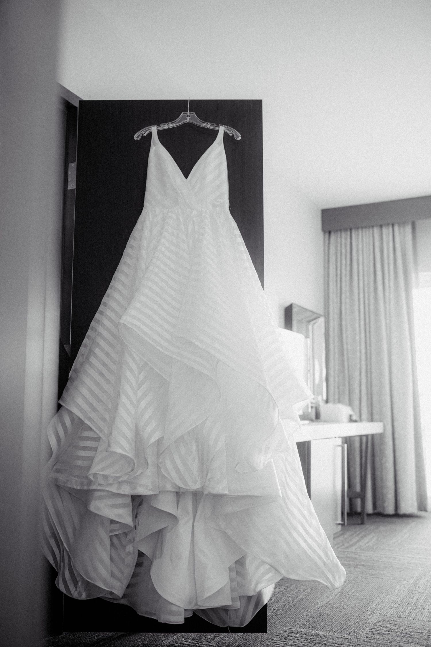 Black and white photo of a ruffled ball gown wedding dress hanging in a hotel room window.