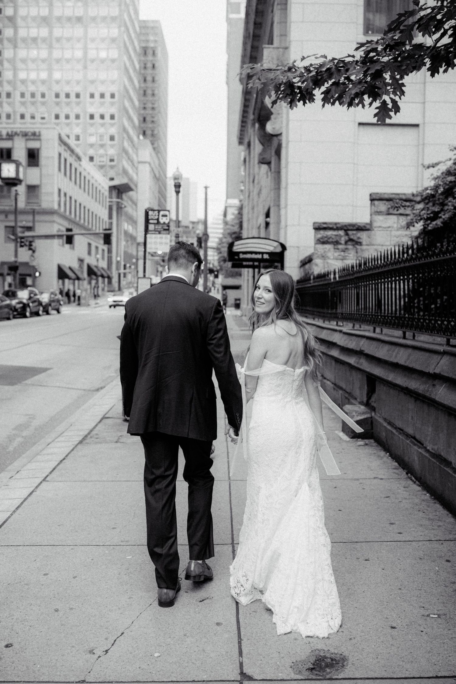 Bride and groom walk hand in hand down a city sidewalk in black and white photo.