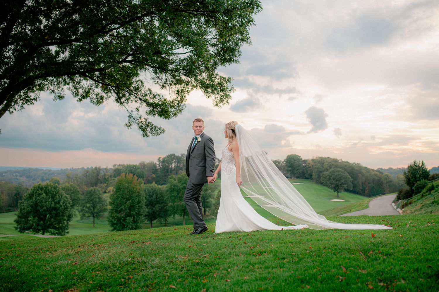 Bride and groom holding hands with dramatic cathedral veil flowing under a large tree outdoors.