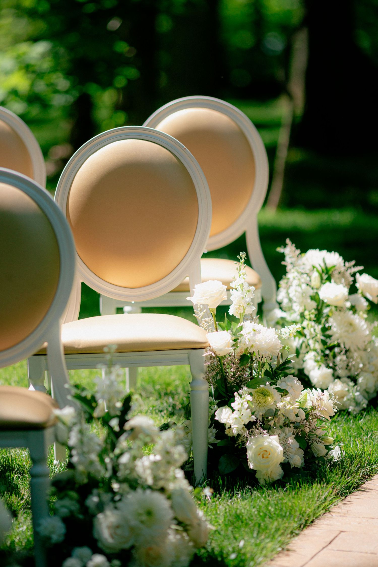 Elegant outdoor wedding ceremony aisle with gold oval-back chairs lined with white floral arrangements.