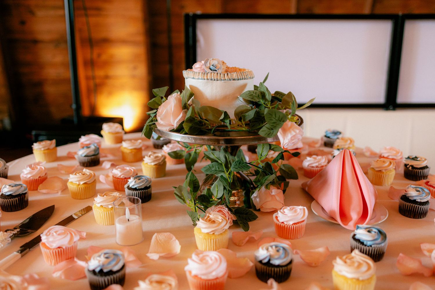 Wedding dessert table with colorful cupcakes surrounding a small cake with floral greenery centerpiece.