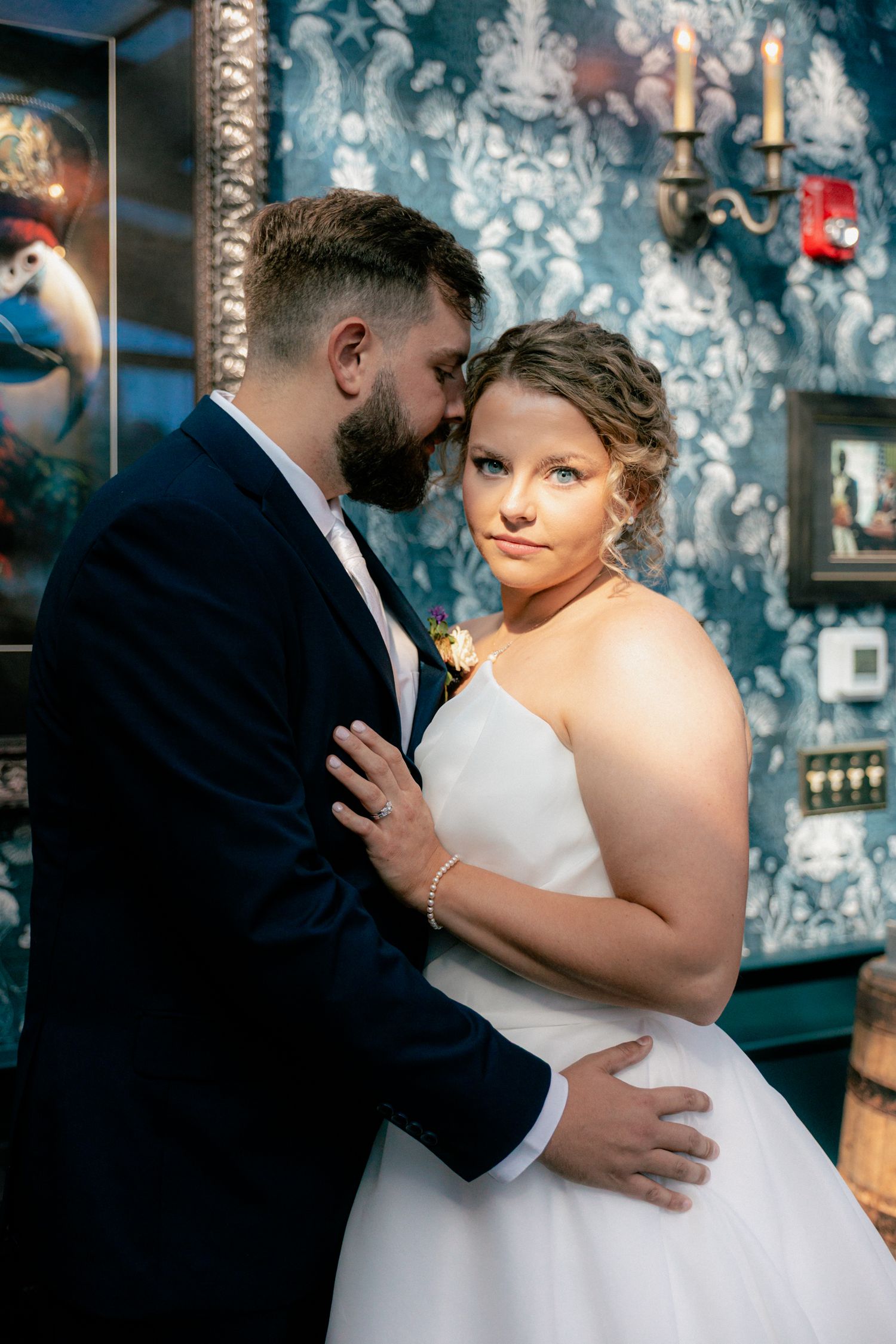 Bearded groom embracing bride against ornate blue wallpaper background.
