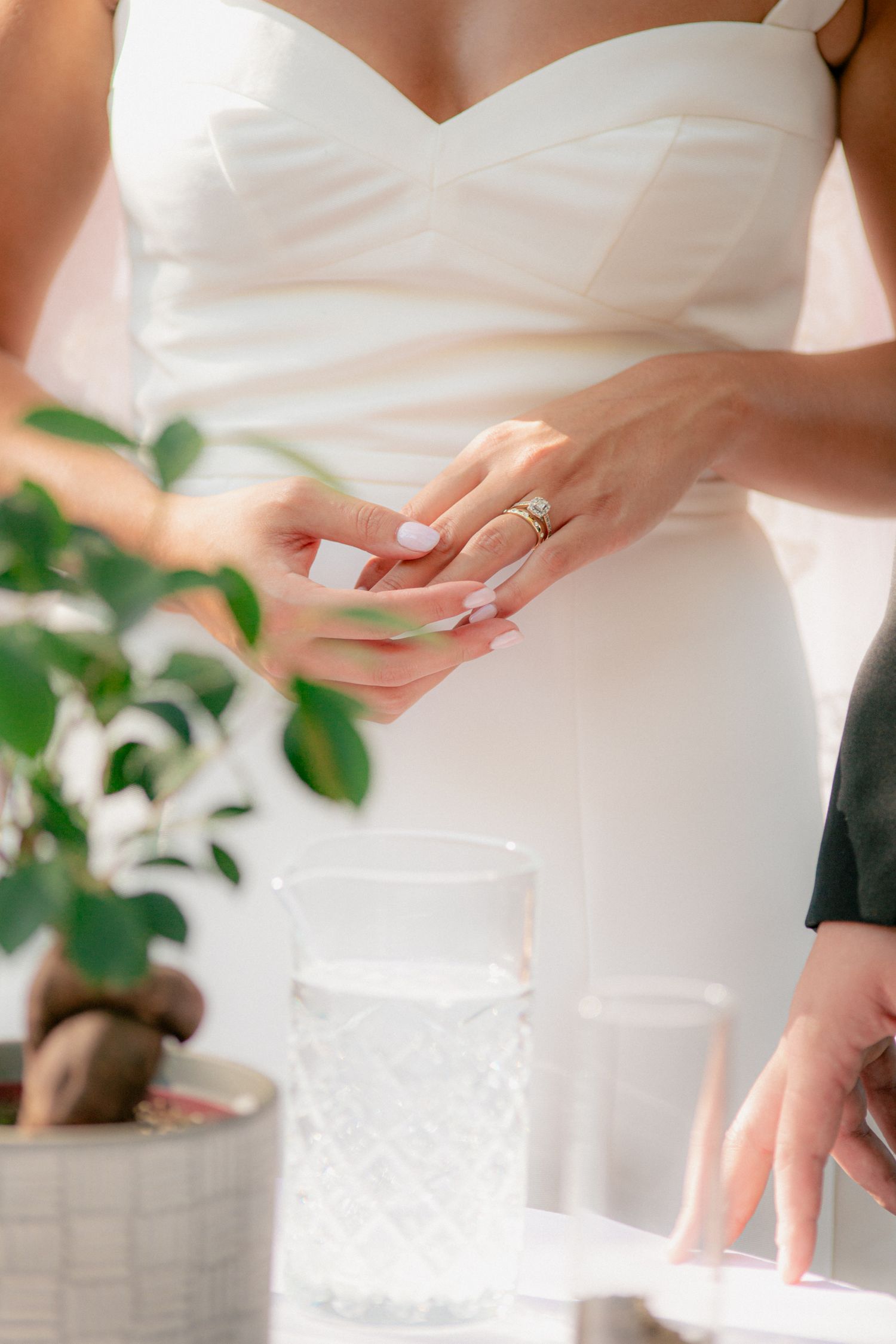 Bride in white dress admiring her diamond wedding ring while standing near a green plant.