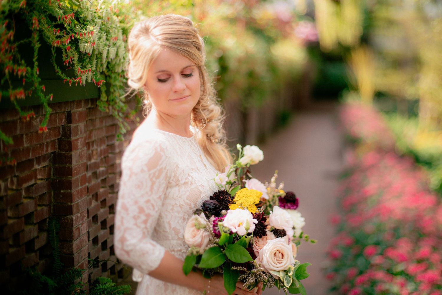 Elegant blonde bride in lace dress admires her colorful bridal bouquet outdoors.