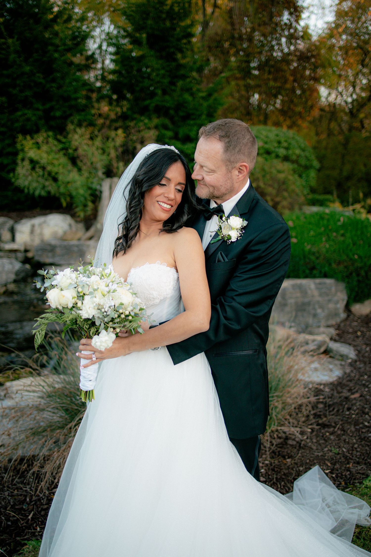 Groom in black tuxedo tenderly embraces smiling dark-haired bride holding white bouquet in an elegant outdoor garden setting.