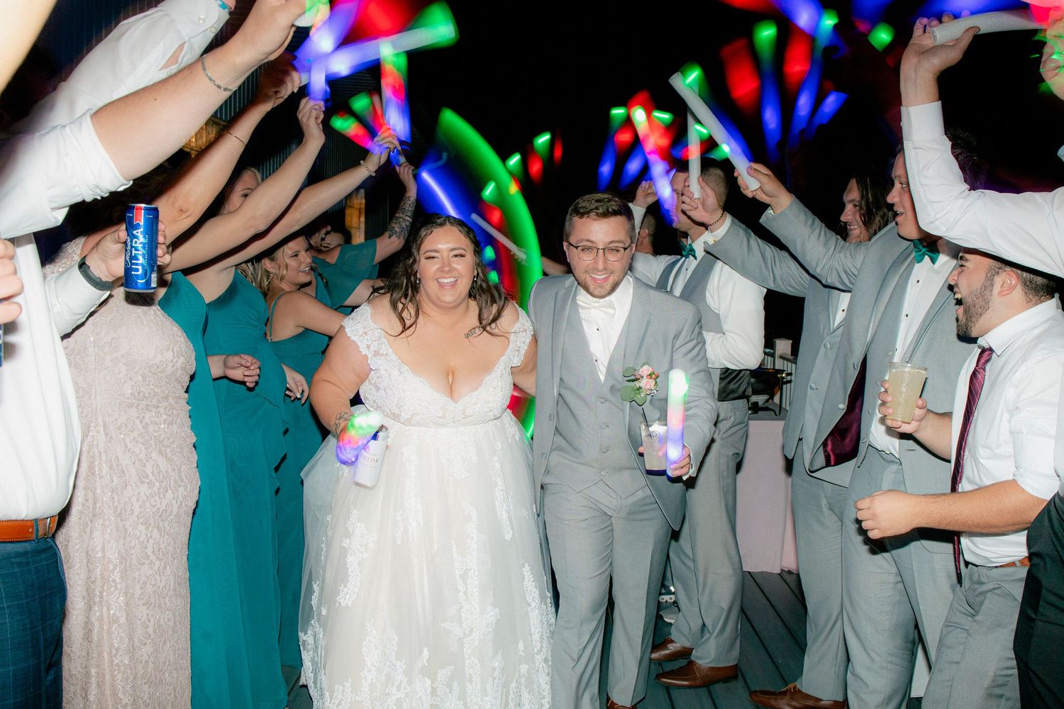 Bride and groom exit wedding reception through tunnel of glowing neon light sticks held by cheering guests.