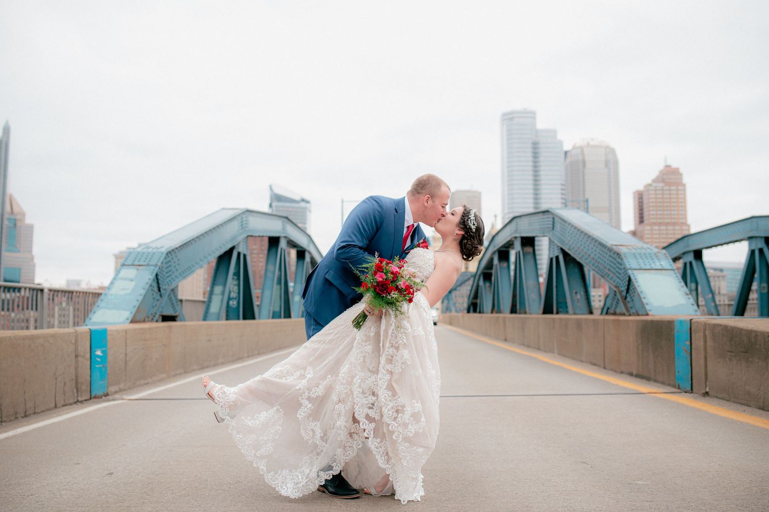 Groom dips bride in lace gown for romantic kiss on Pittsburgh bridge with city skyline behind them.