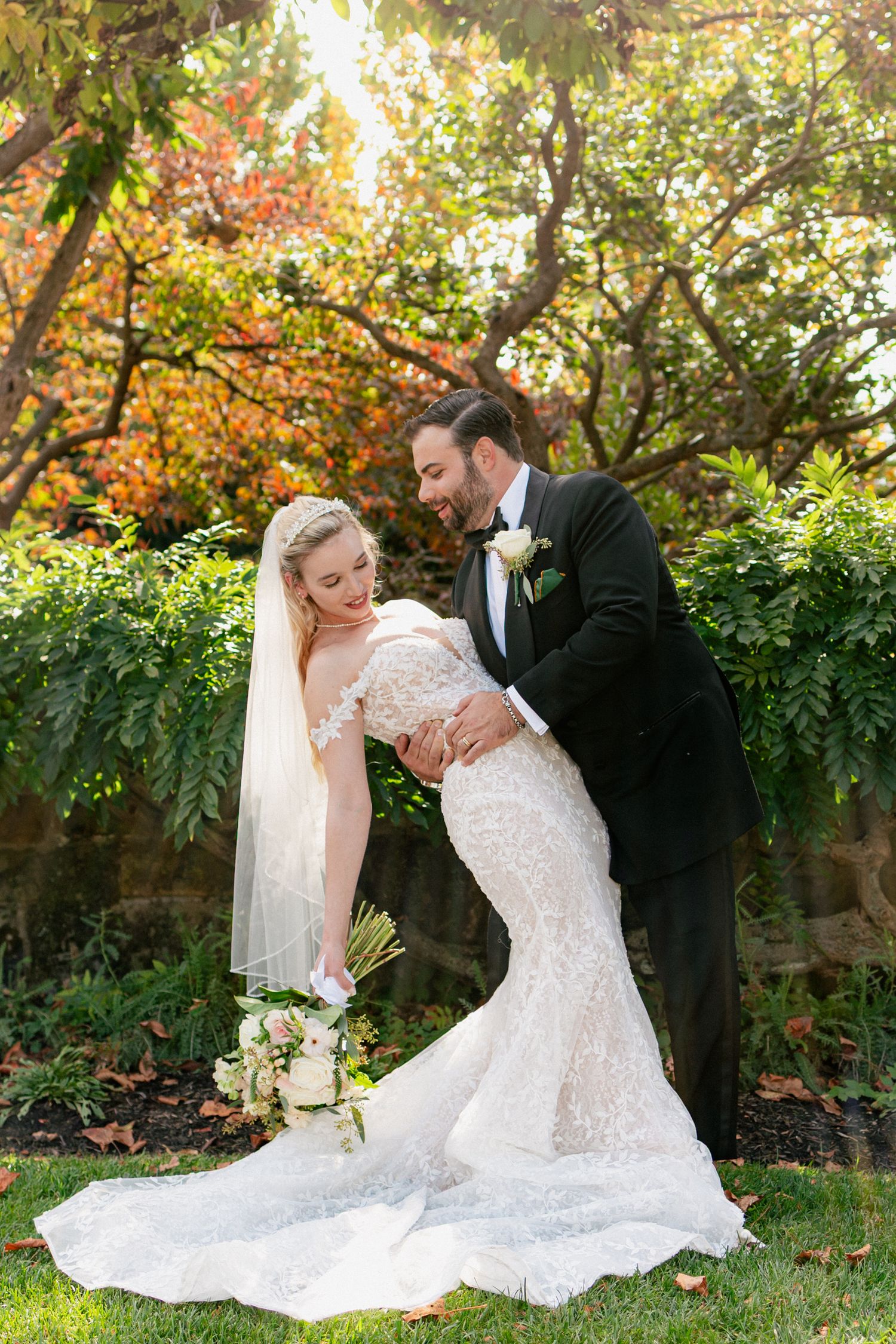 Groom in black tuxedo playfully dips laughing bride in lace mermaid gown amid autumn foliage.