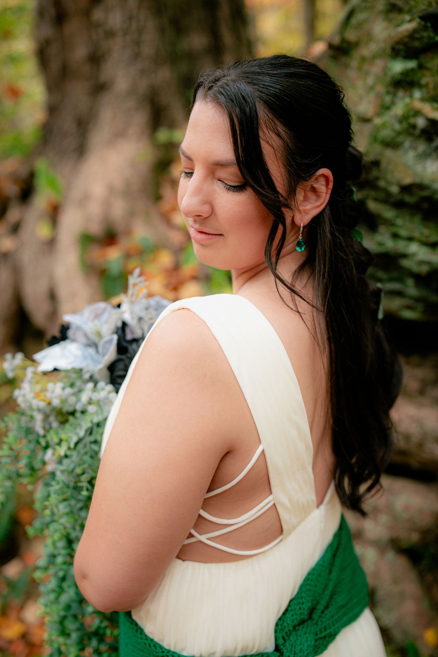 Bride in white gown with criss-cross back detail holds bouquet while posing in autumn woodland setting.