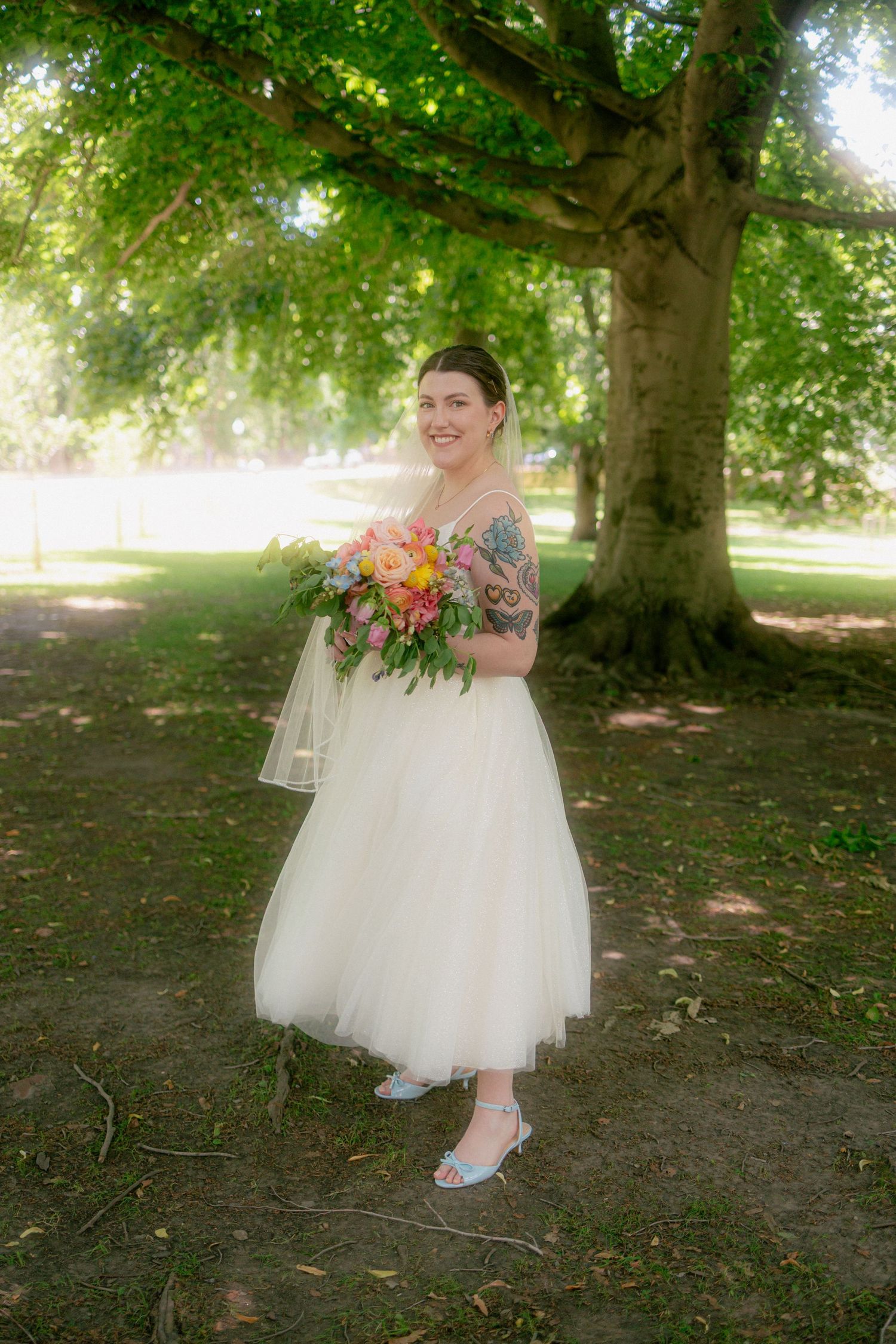 Bride in white tulle midi dress holding colorful bouquet, standing under large tree in sunny park, with tattoos and blue heels.