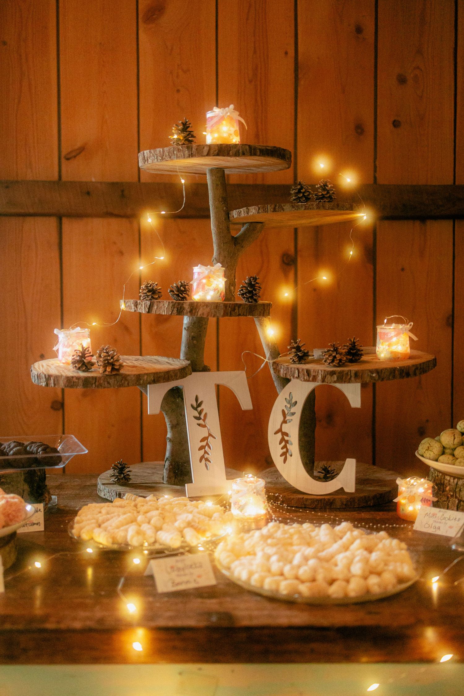 Rustic wedding dessert table with tiered wood slab stand, pine cones, fairy lights, and white candy dishes in a barn setting.