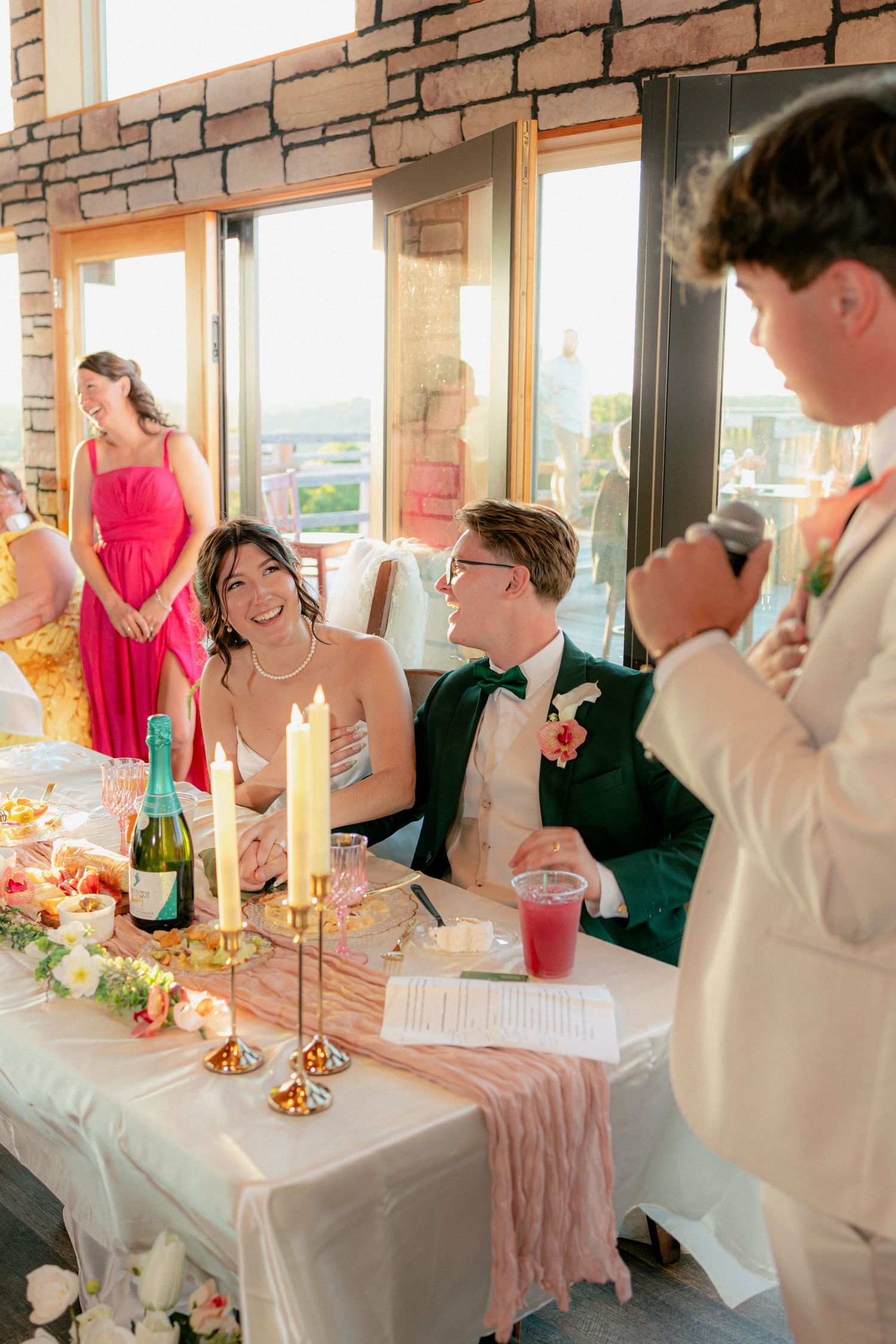 Bride and groom laugh at sweetheart table during wedding reception toast, with candles and champagne nearby.