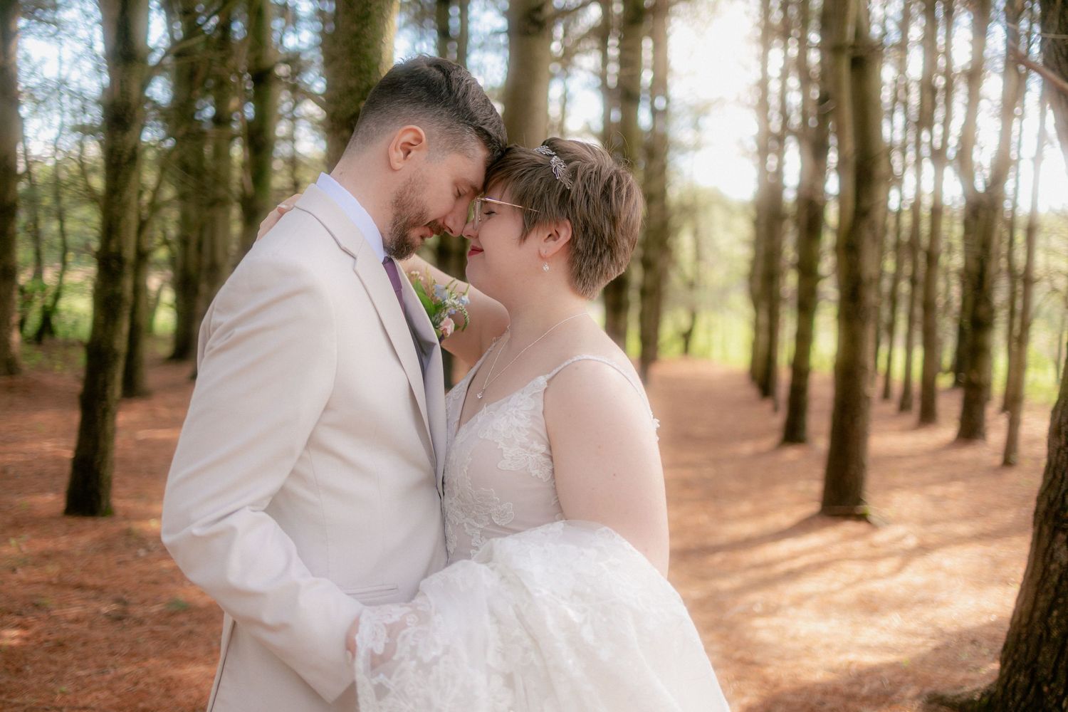 Bride and groom share a romantic forehead touch in a sunlit pine forest, bride in lace gown, groom in beige suit.
