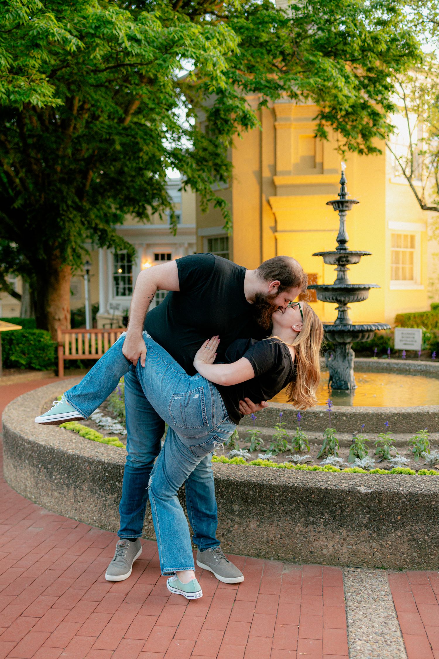 Couple sharing a dip kiss in front of a stone fountain surrounded by lush green trees.