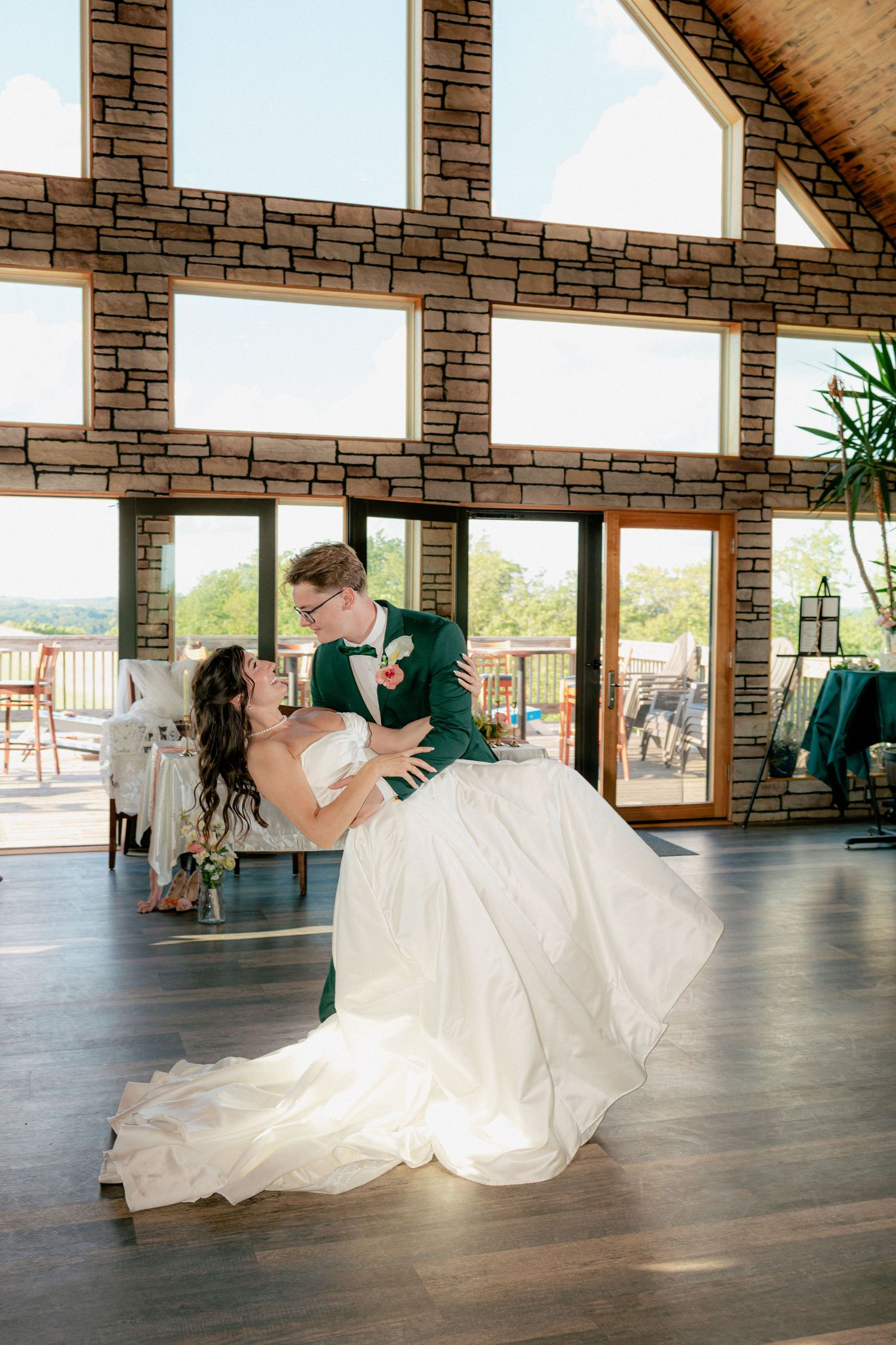Groom in green suit dips bride in white gown during first dance in rustic stone venue with large windows.