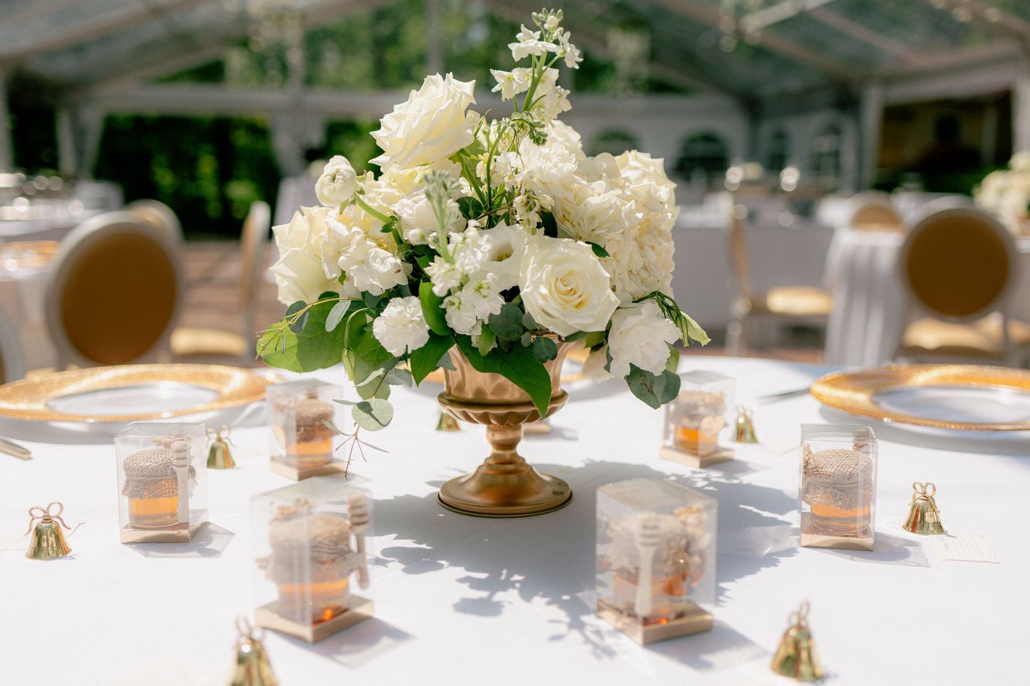 Gold urn centerpiece with white roses and greenery on a white tablecloth with gold favor boxes.