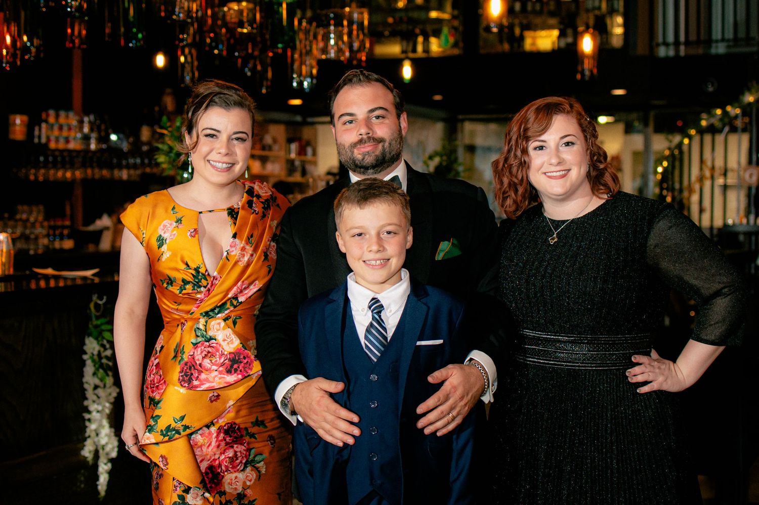 Family of four poses together at wedding reception, featuring two adult sisters, a young boy in navy suit, and the groom.