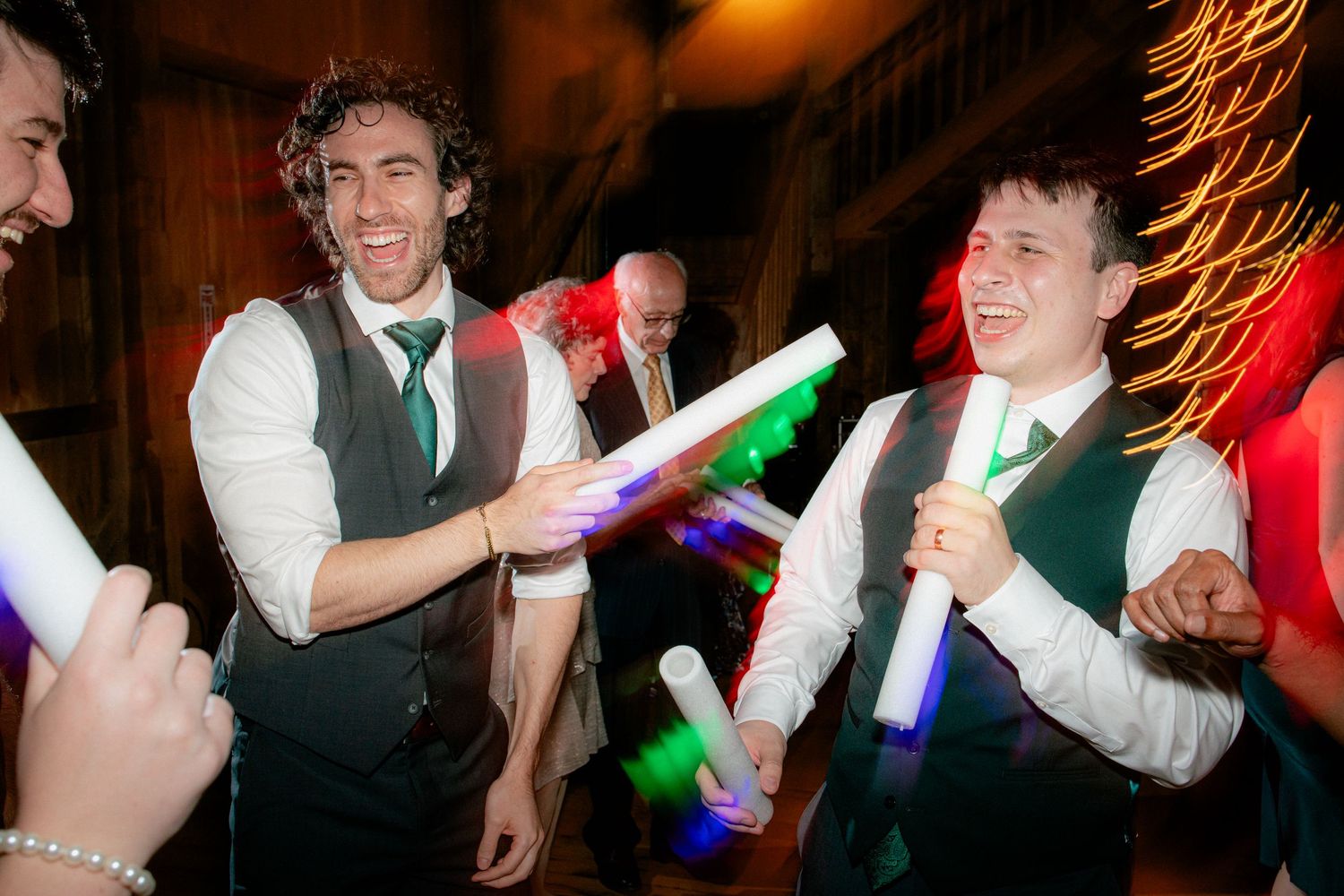 Groomsmen laugh and dance with colorful glow sticks during a festive wedding reception celebration.