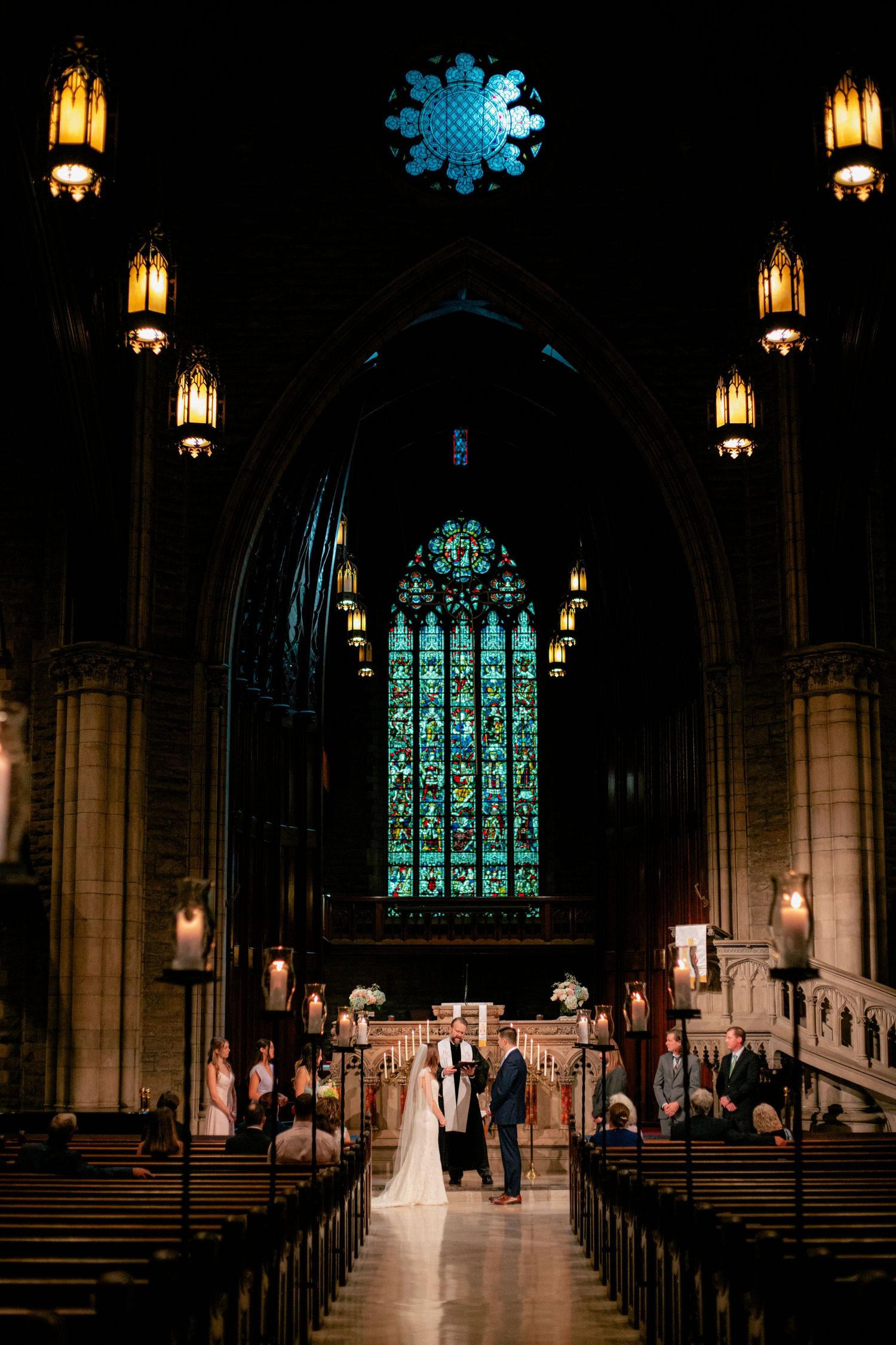 Bride and groom stand at altar of stunning gothic cathedral with glowing stained glass window backdrop.