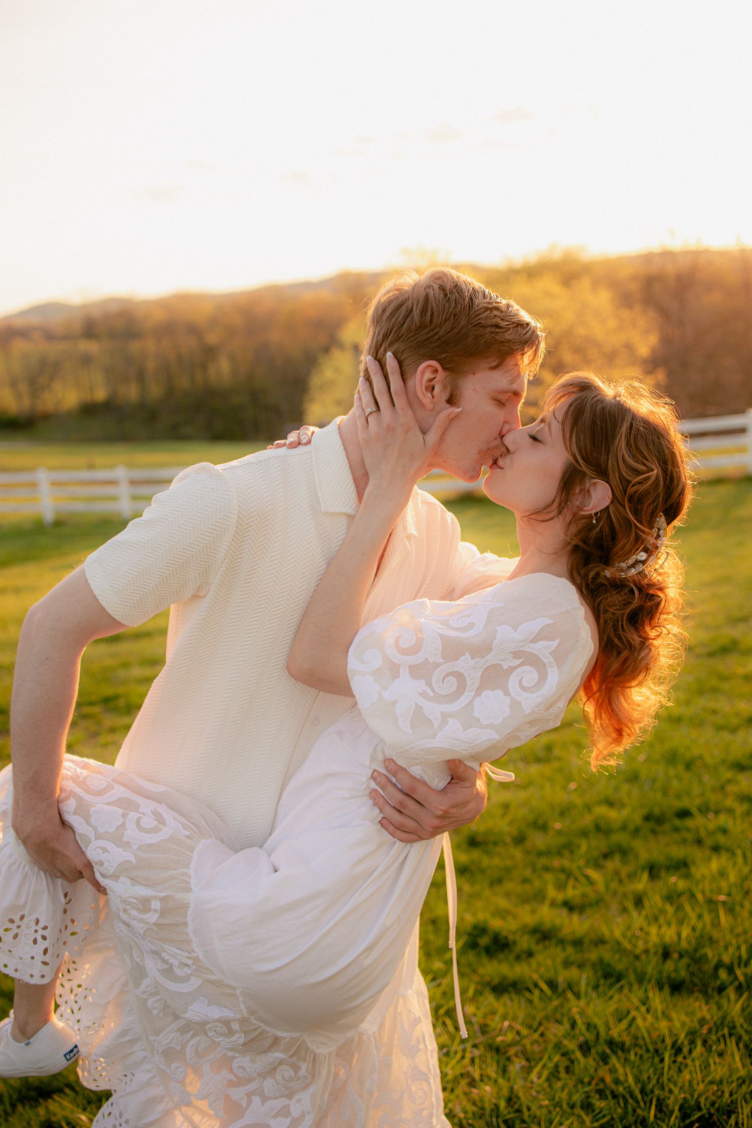 Groom dips and kisses bride in white lace dress at golden hour on a lush green farm field.