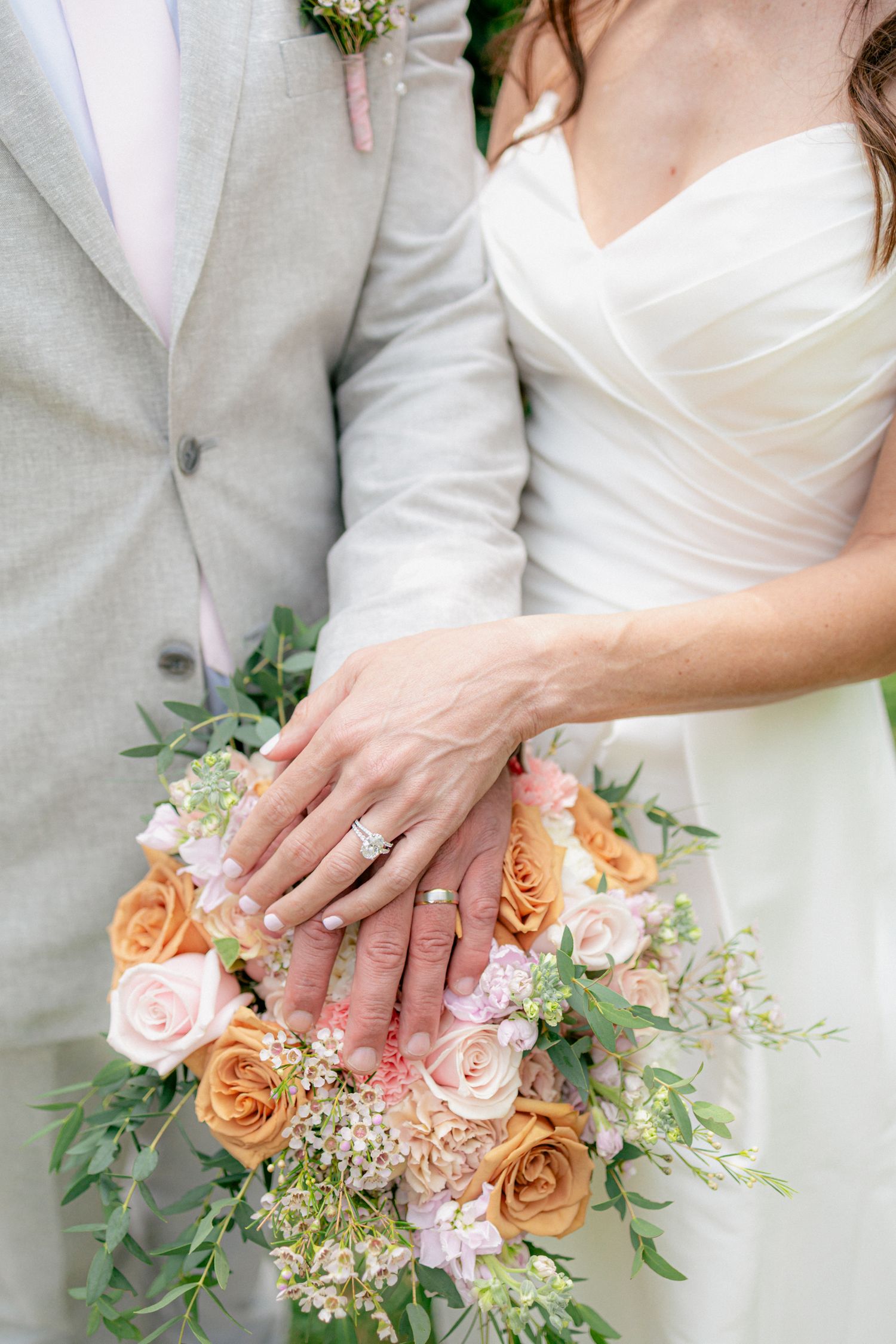 Close-up of bride and groom placing hands on a peach and blush rose bouquet, showcasing wedding rings.
