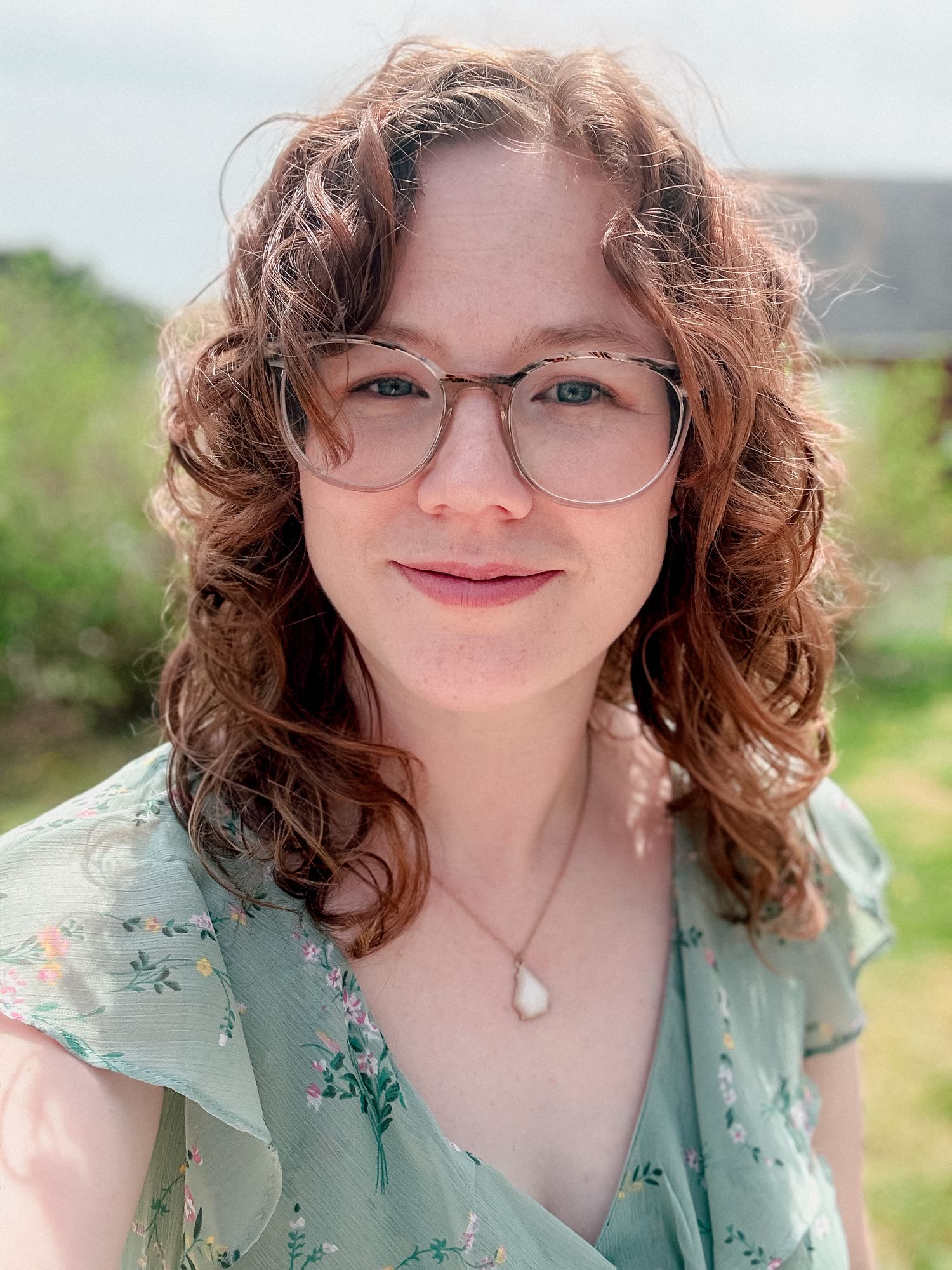 Young woman with curly auburn hair and glasses smiles outdoors in floral green dress on sunny day.