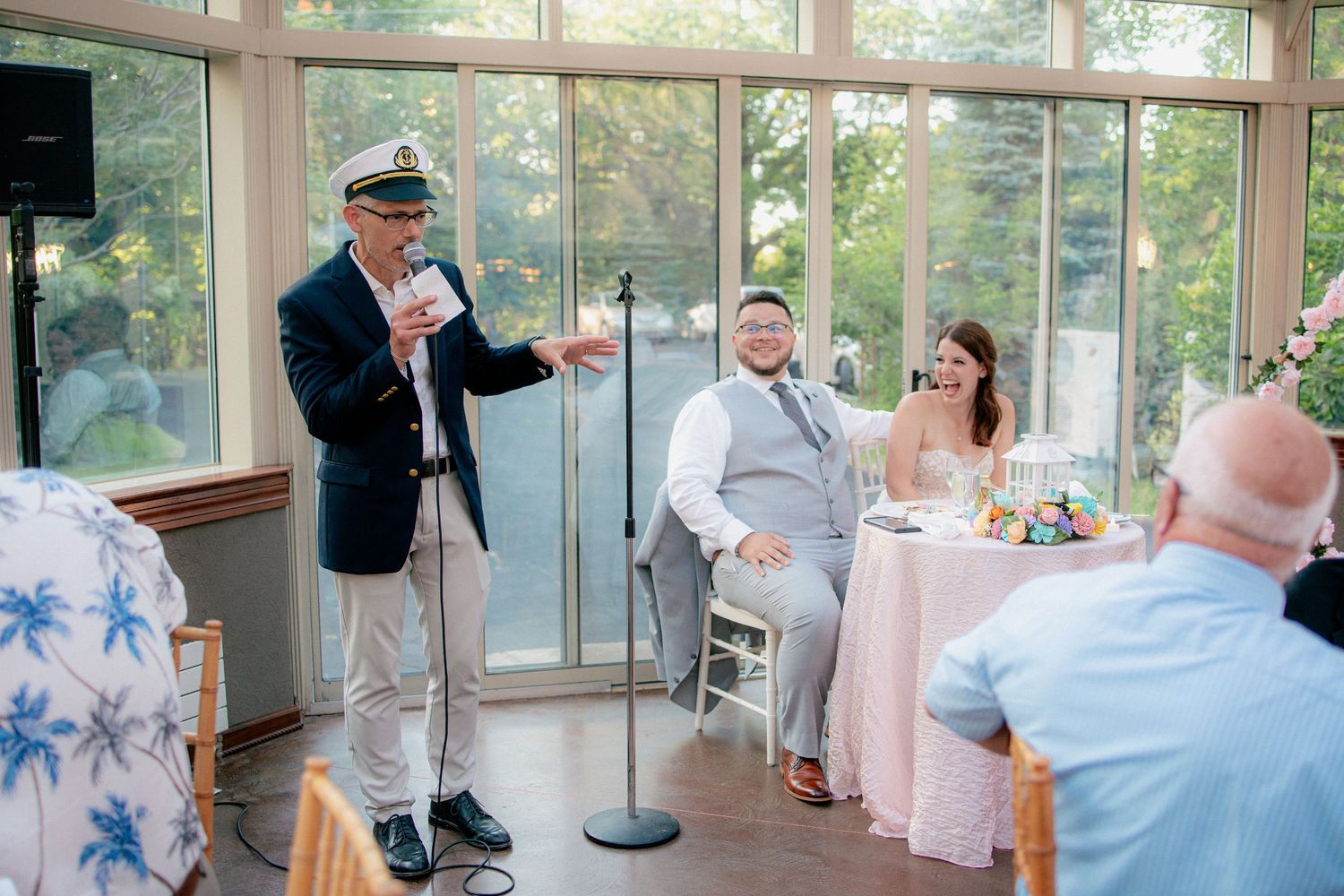 Man in captain hat giving speech at microphone while bride and groom smile at wedding reception.