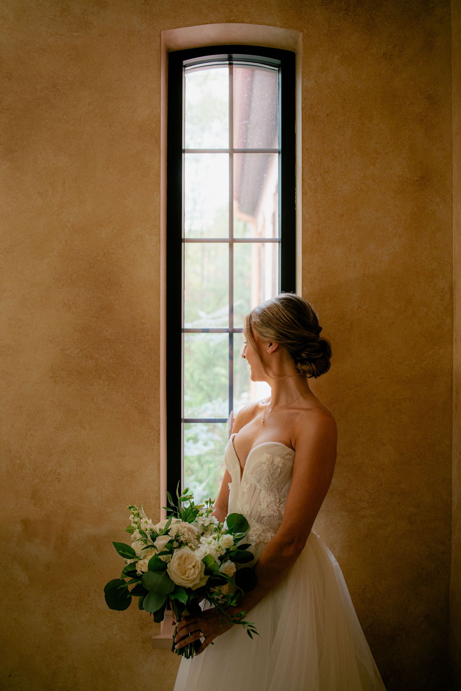Elegant bride in strapless gown holds white bouquet while gazing through tall arched window in warmly lit stone room.