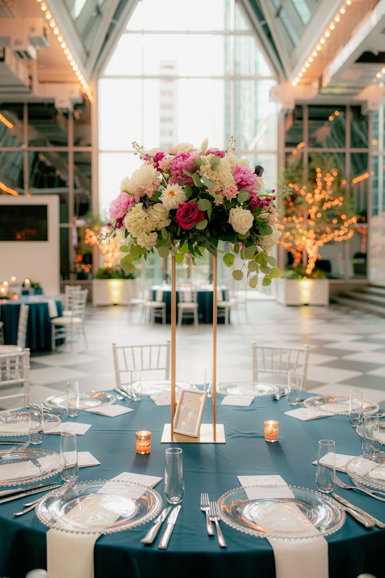 Elegant round wedding table with tall gold-stand floral centerpiece of pink and white blooms, navy linen, and crystal chargers.