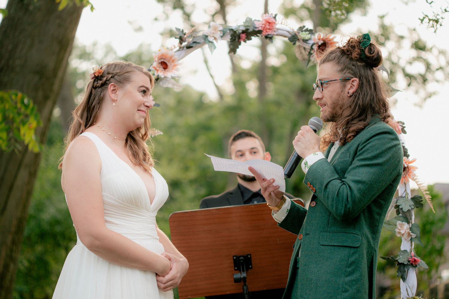 Groom in emerald green suit reads vows to bride at romantic outdoor garden wedding ceremony.