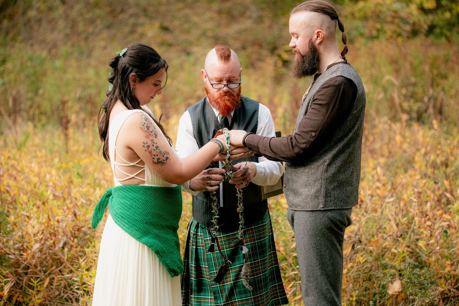 Bride and groom participate in Celtic handfasting ceremony with officiant in kilt outdoors.