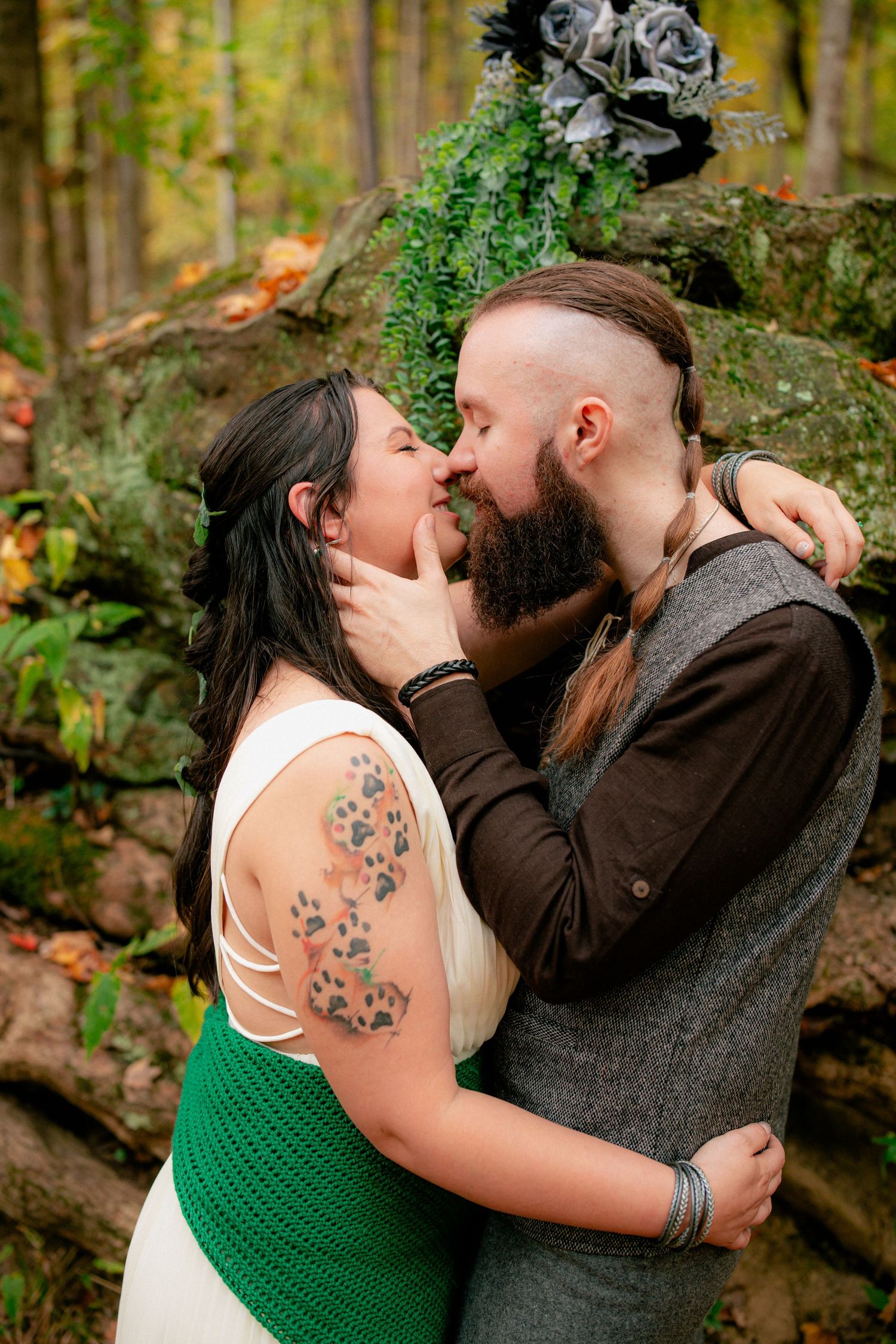 Couple shares a romantic kiss near rock wall decorated with dark floral arrangement in fall forest.
