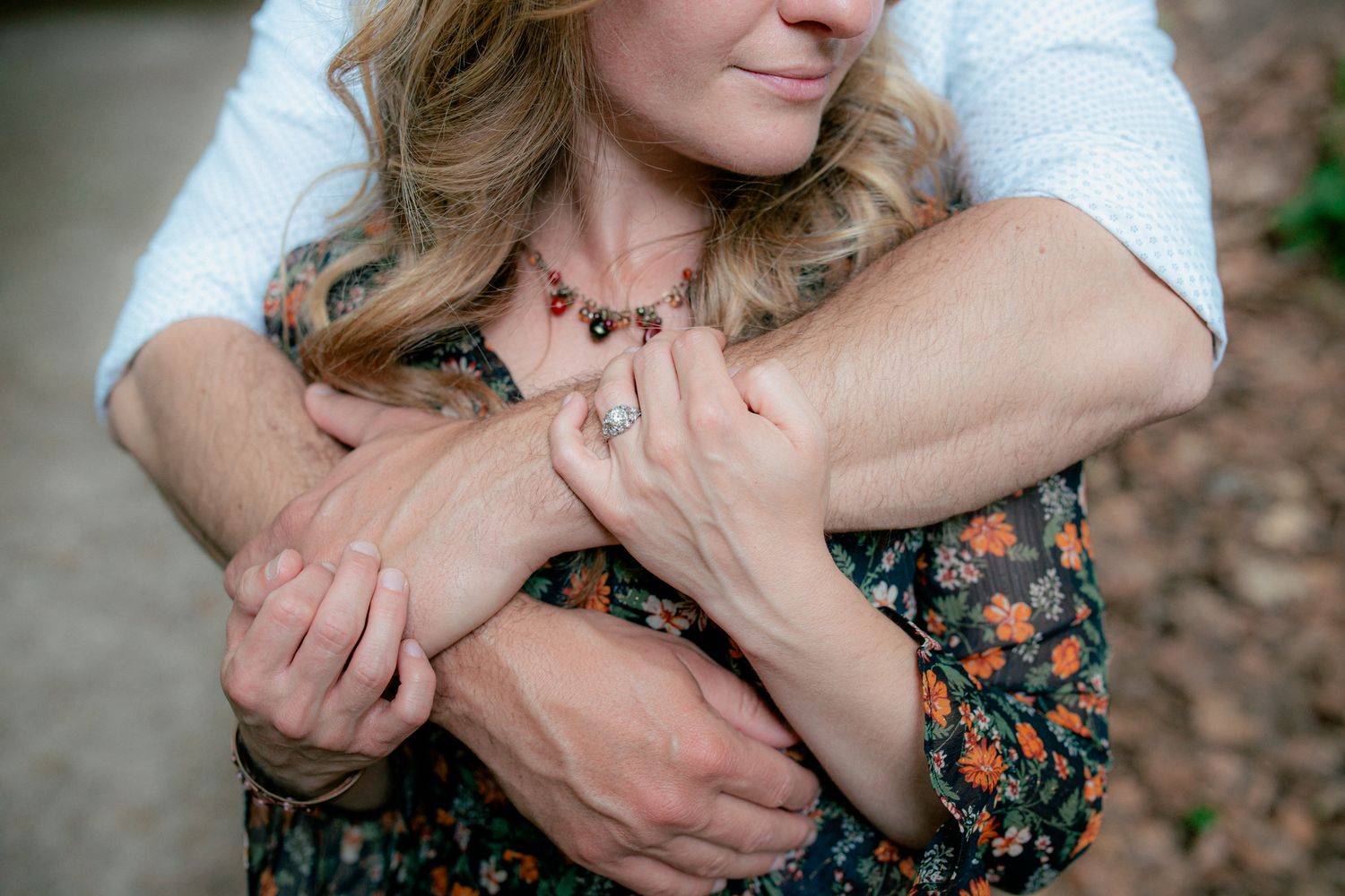 Close-up of couple embracing from behind, hands intertwined, woman in floral dress outdoors.