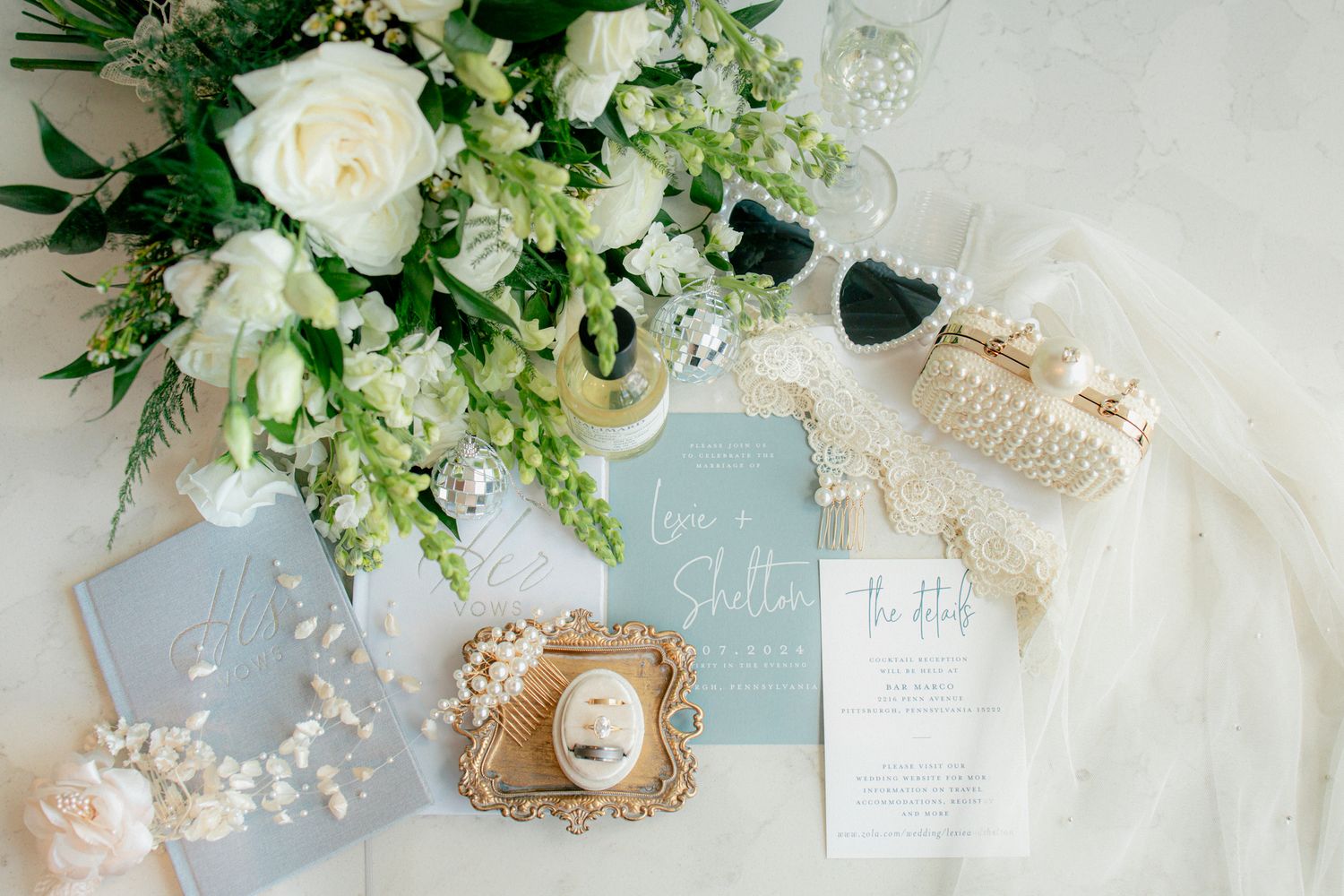 Elegant wedding flat lay featuring white floral bouquet, dusty blue stationery, pearl accessories, and heart sunglasses.