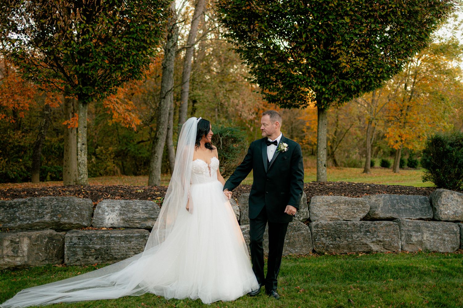 Bride and groom holding hands in a lush autumn park, bride wearing a long white veil and gown.