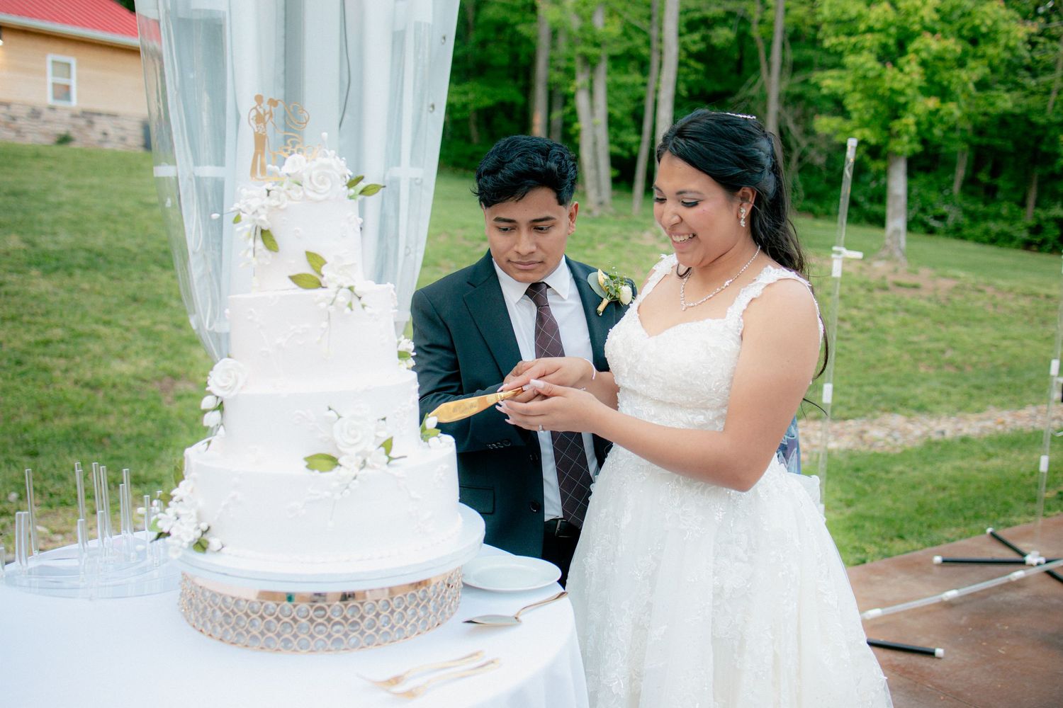 Bride and groom cutting elegant four-tier white wedding cake decorated with greenery outdoors.