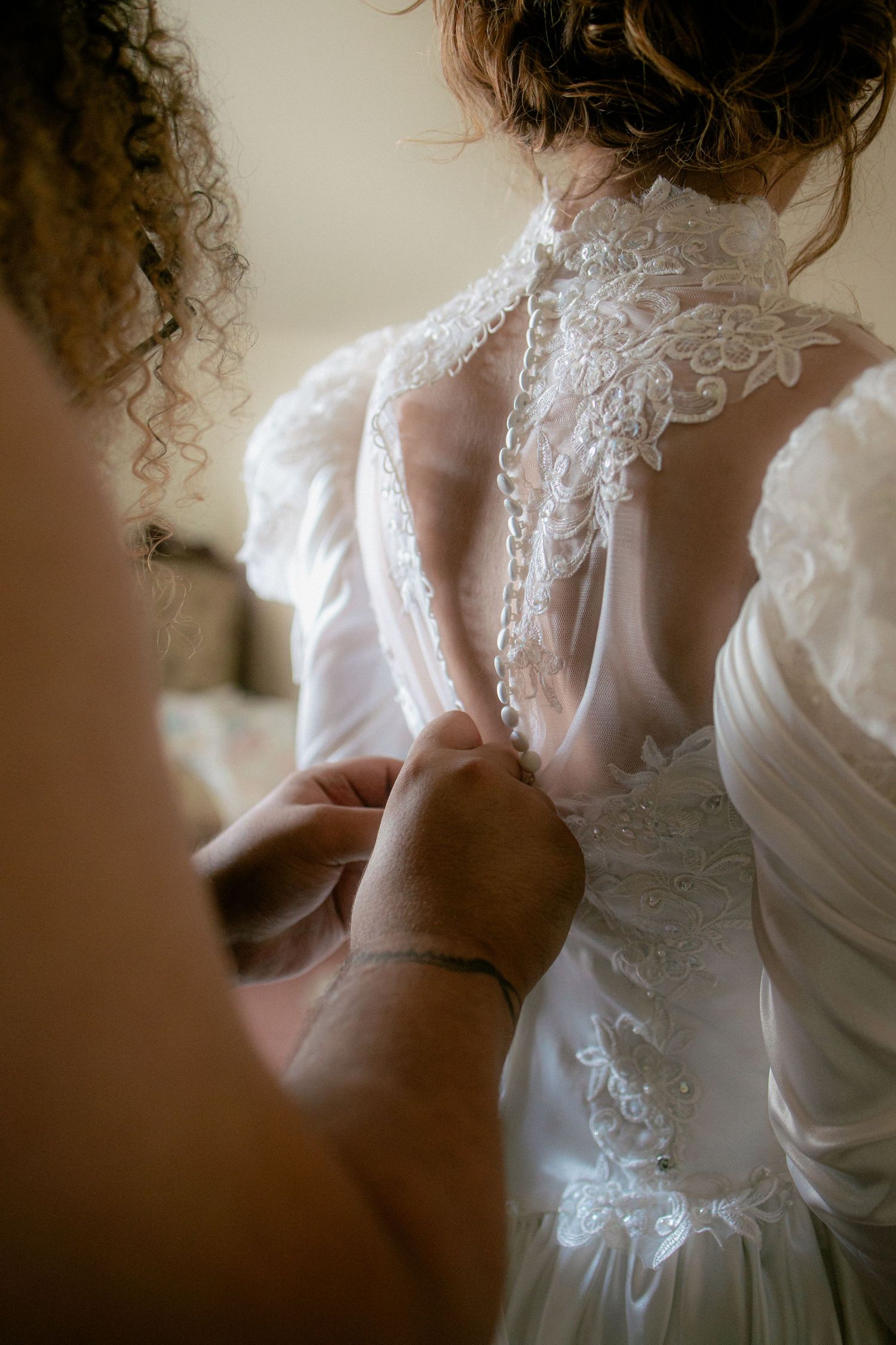 Close-up of hands buttoning the intricate lace back of a vintage white wedding dress.
