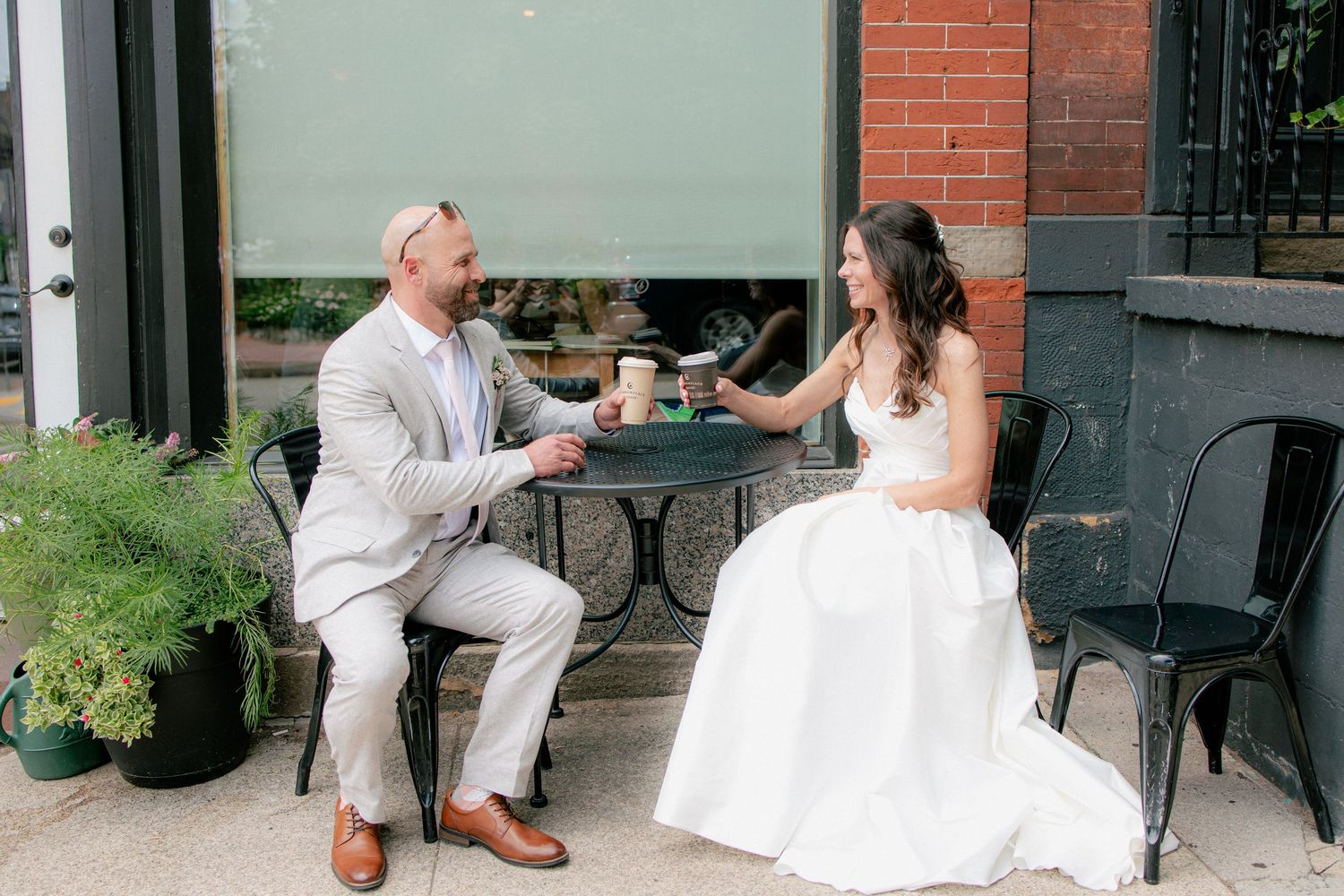 Bride and groom share a relaxed coffee date moment at an outdoor cafe table on their wedding day.