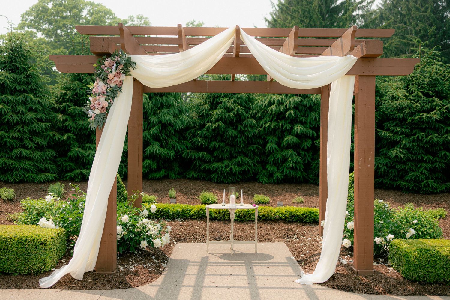 Wooden pergola wedding arch draped with white fabric and pink flowers in a garden ceremony setting.