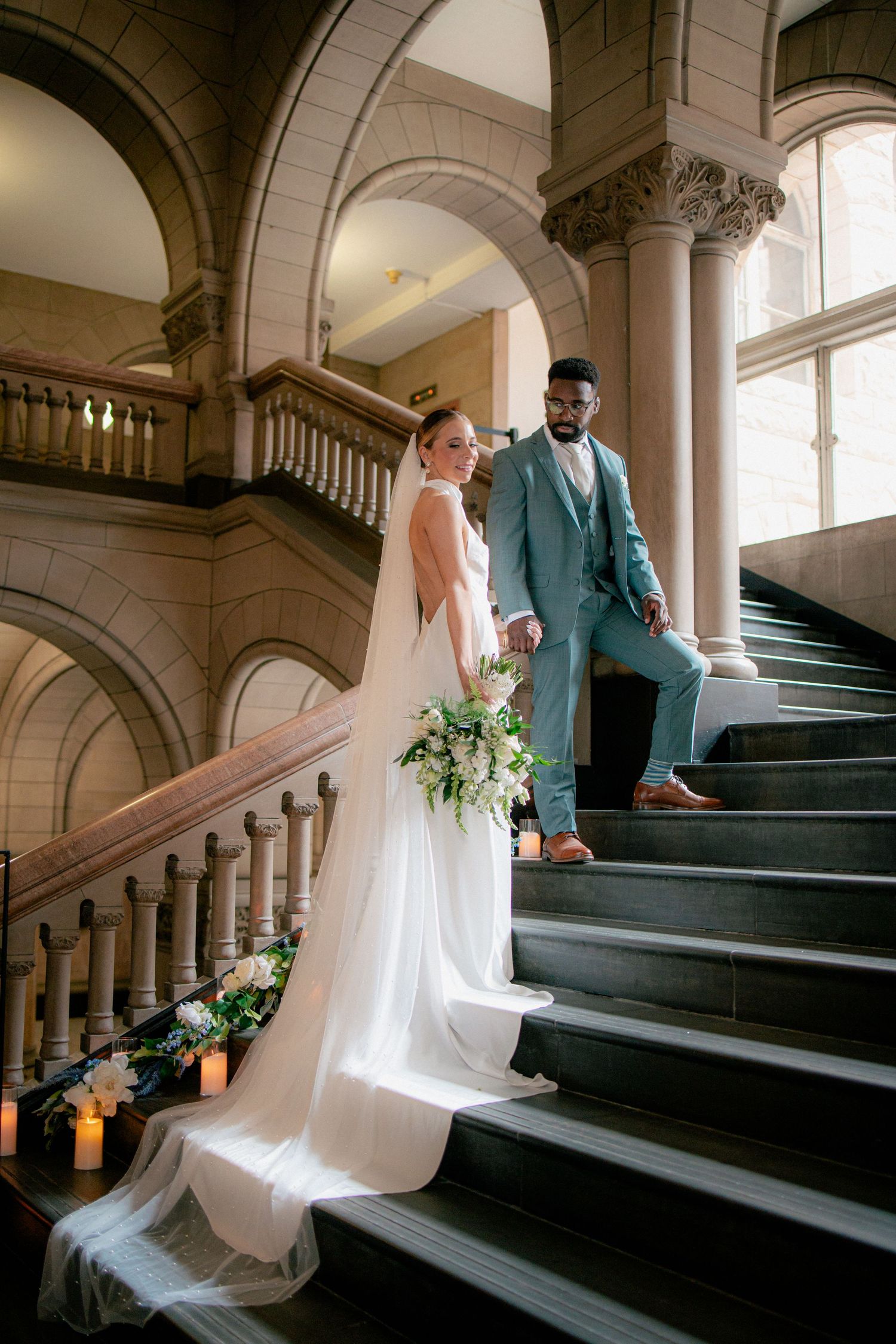 Bride holds white floral bouquet on candlelit stone staircase as groom in teal suit stands beside her.