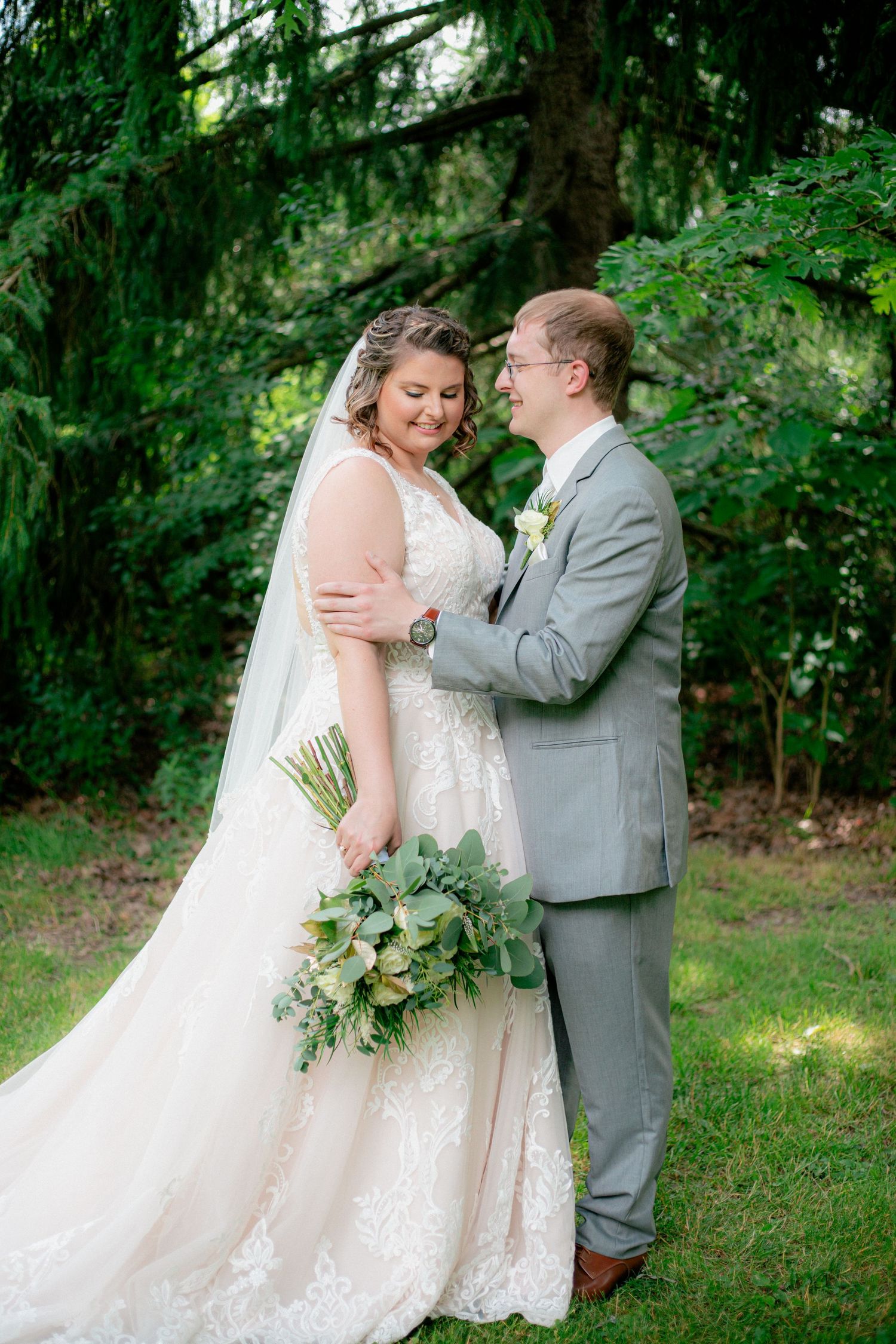 Bride in lace gown holding greenery bouquet smiling with groom in gray suit outdoors.