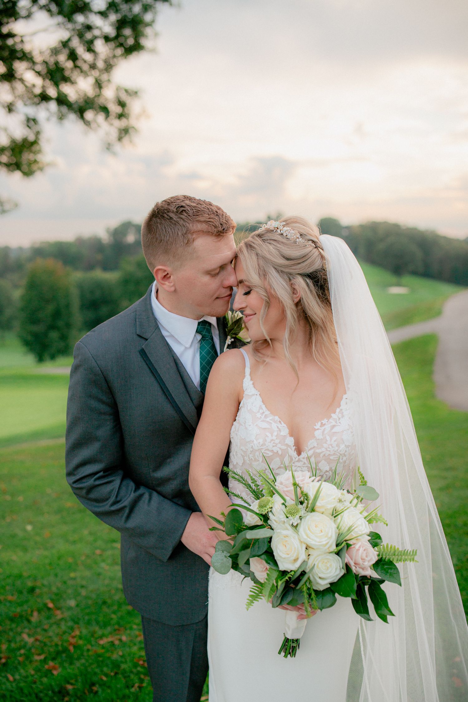 Bride holding white rose bouquet as groom nuzzles her cheek at sunset on a green hillside.
