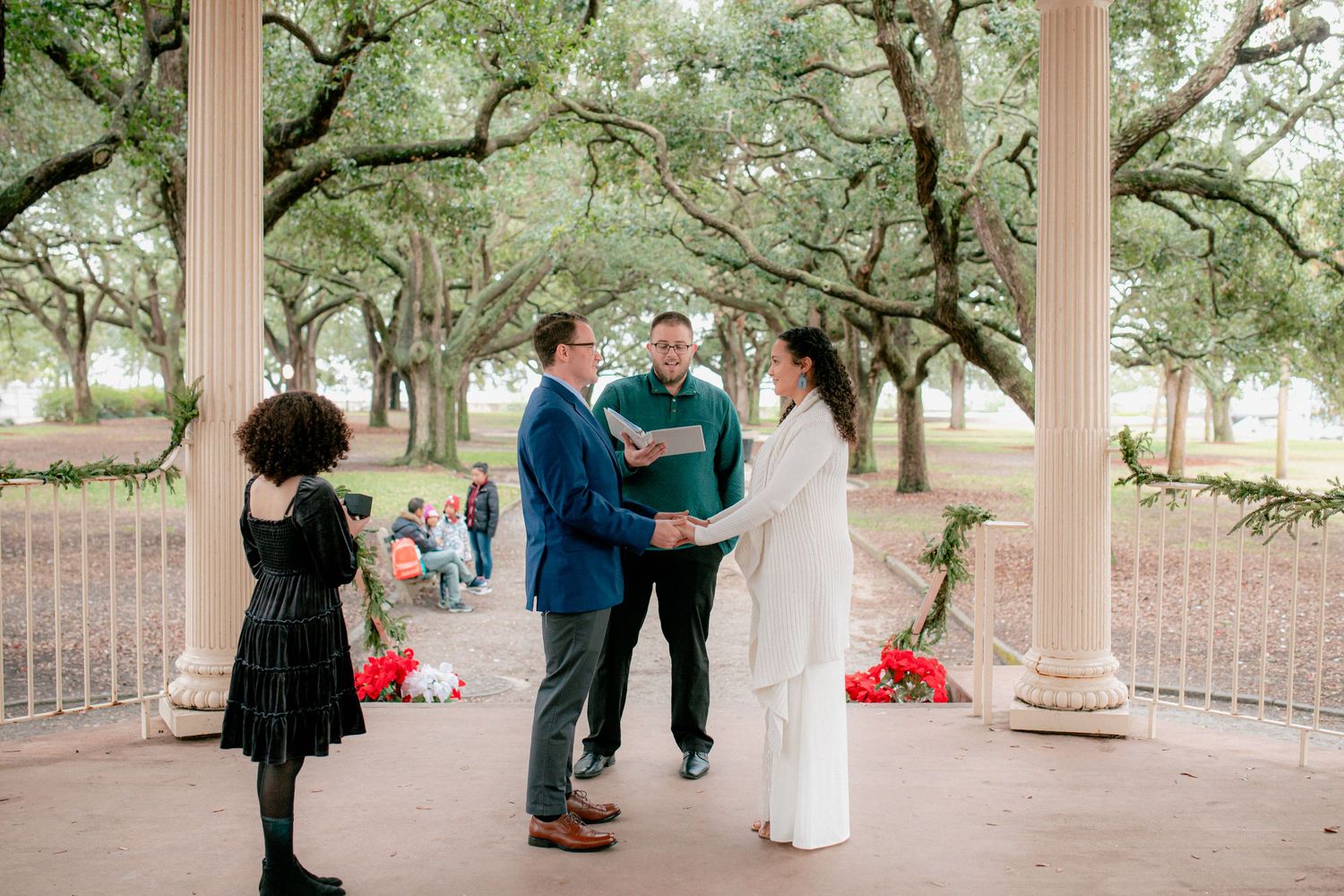 Couple holds hands during outdoor wedding ceremony under oak trees at historic columned pavilion with small gathering.