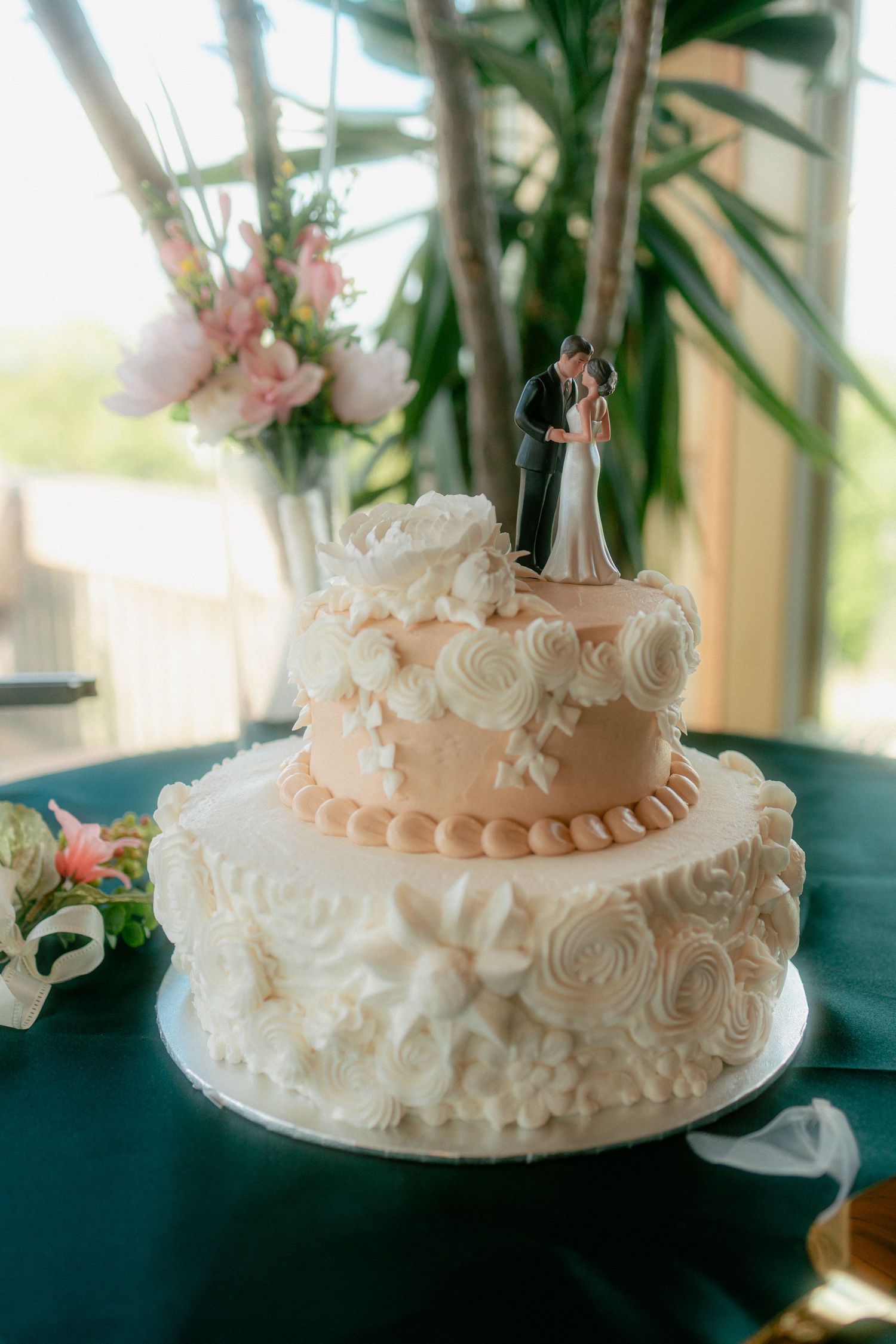 Two-tier wedding cake with bride and groom topper, white rosette frosting, and peach accents on a teal tablecloth.