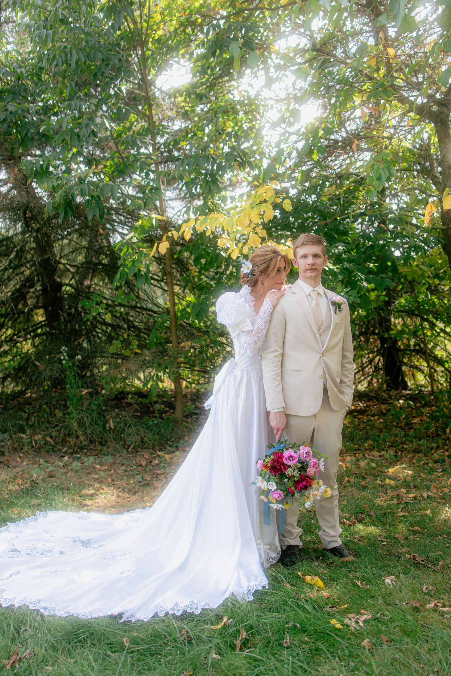 Bride in vintage lace gown with long train and groom in beige suit pose under lush green trees.