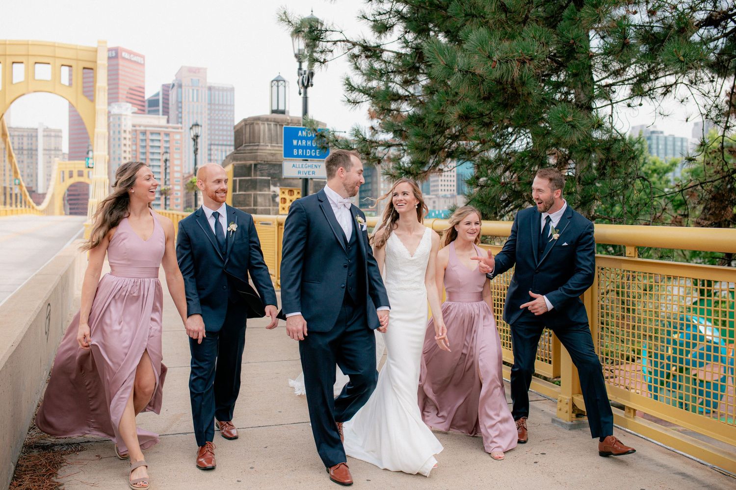 Wedding party walks on Pittsburgh bridge, bridesmaids in dusty rose gowns, groomsmen in navy suits, bride and groom at center.