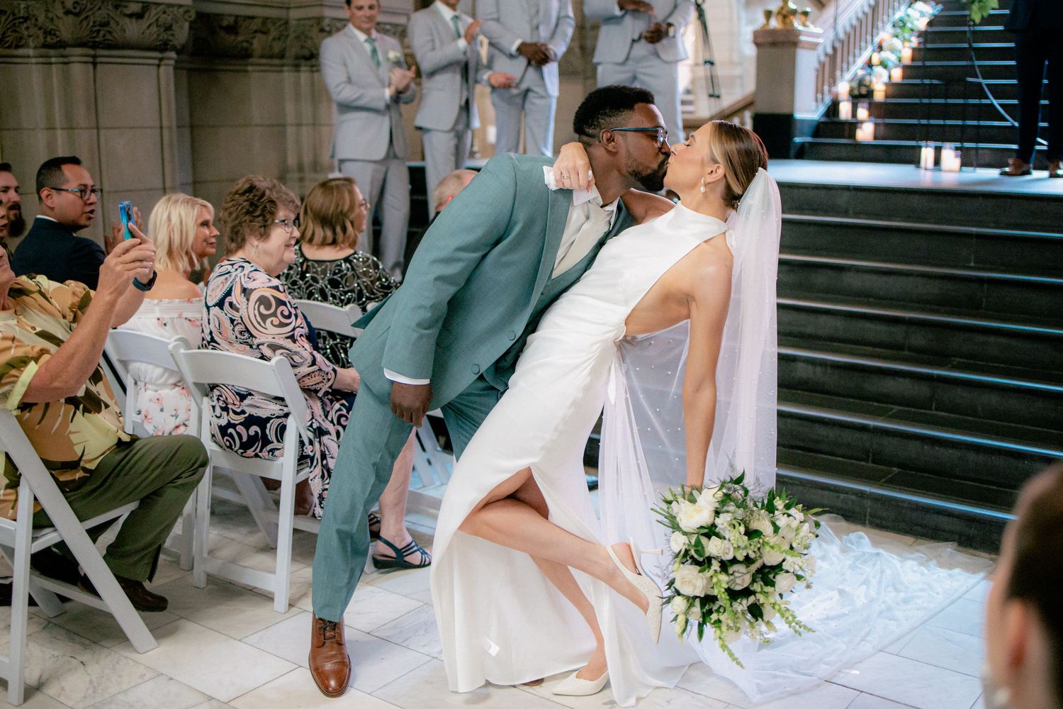 Interracial couple sharing first kiss at wedding ceremony while bride holds white bouquet and lifts leg.